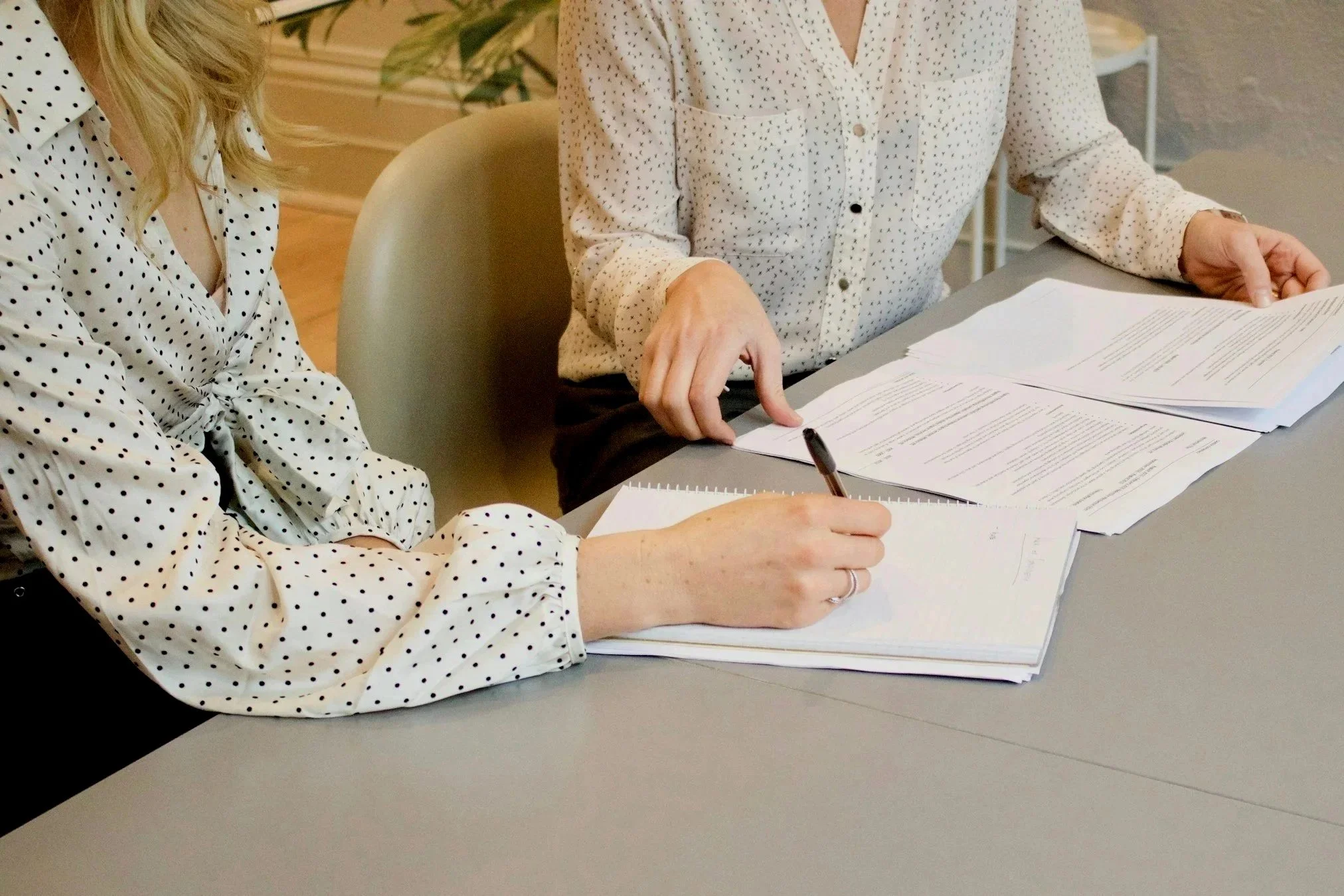 Two women at a desk reviewing and taking notes on documents and papers, dressed in white blouses with polka dots.
