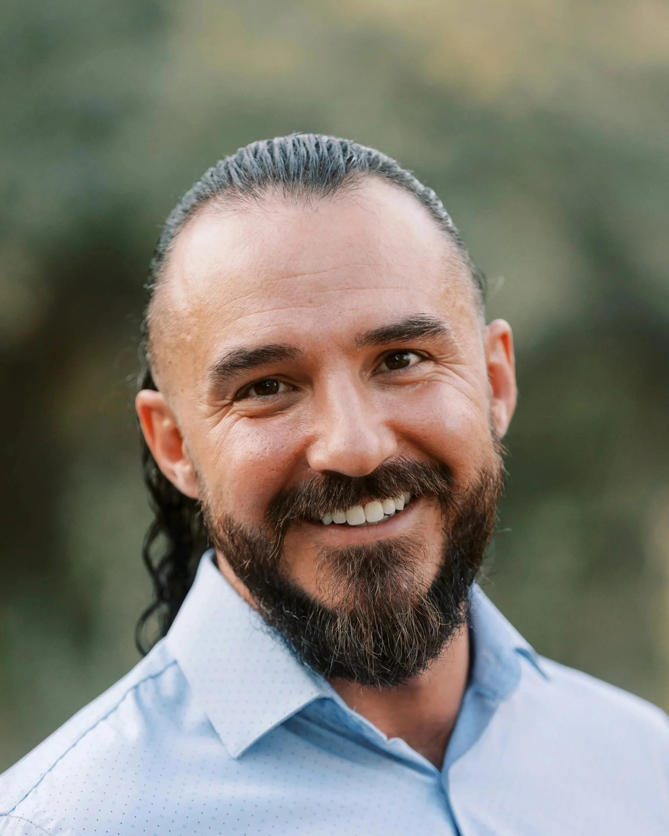 Close-up of a smiling man with dark hair slicked back and a full beard, wearing a light blue collared shirt, outdoors with a blurred natural background.
