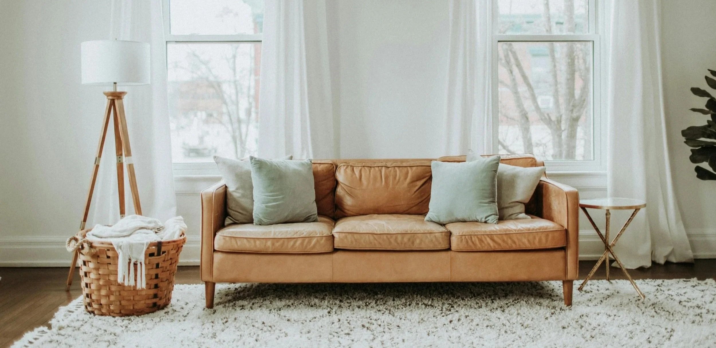 Living room with a tan leather sofa, four light green pillows, two large windows with white curtains, a wooden floor lamp, a woven laundry basket with white towels, a small round wooden side table, and a white shag rug.