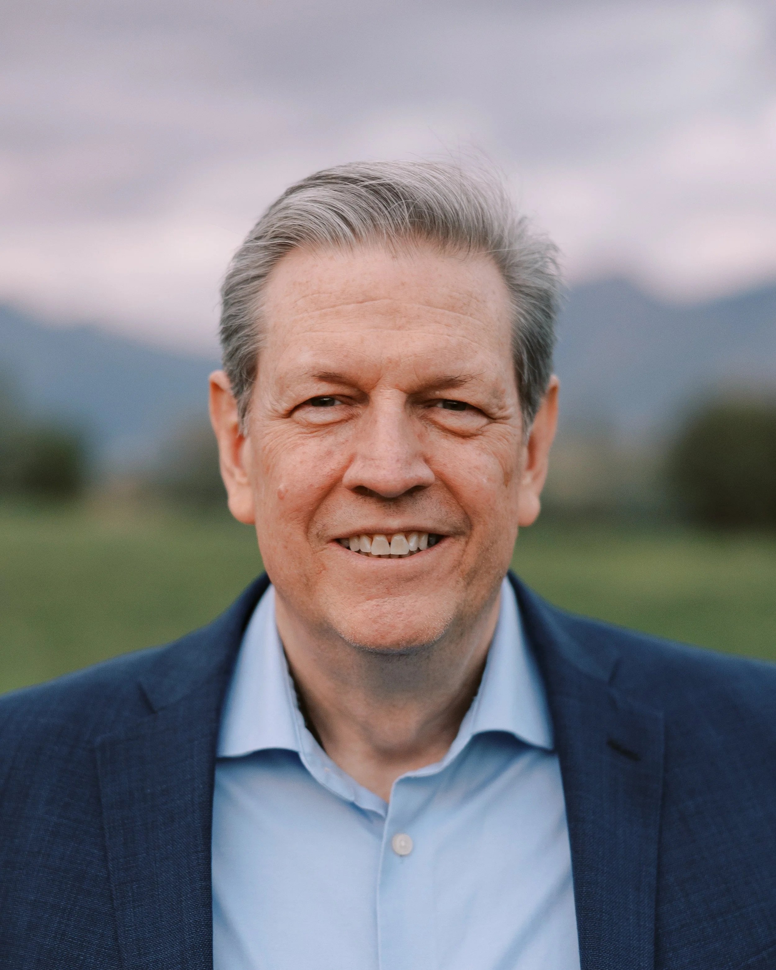 Portrait of a smiling middle-aged man with gray hair, wearing a blue suit jacket and light blue collared shirt, outdoors with mountains and cloudy sky in the background.
