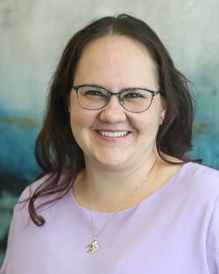 A woman smiling with glasses, wearing a light purple top and a necklace, against a blurred background.