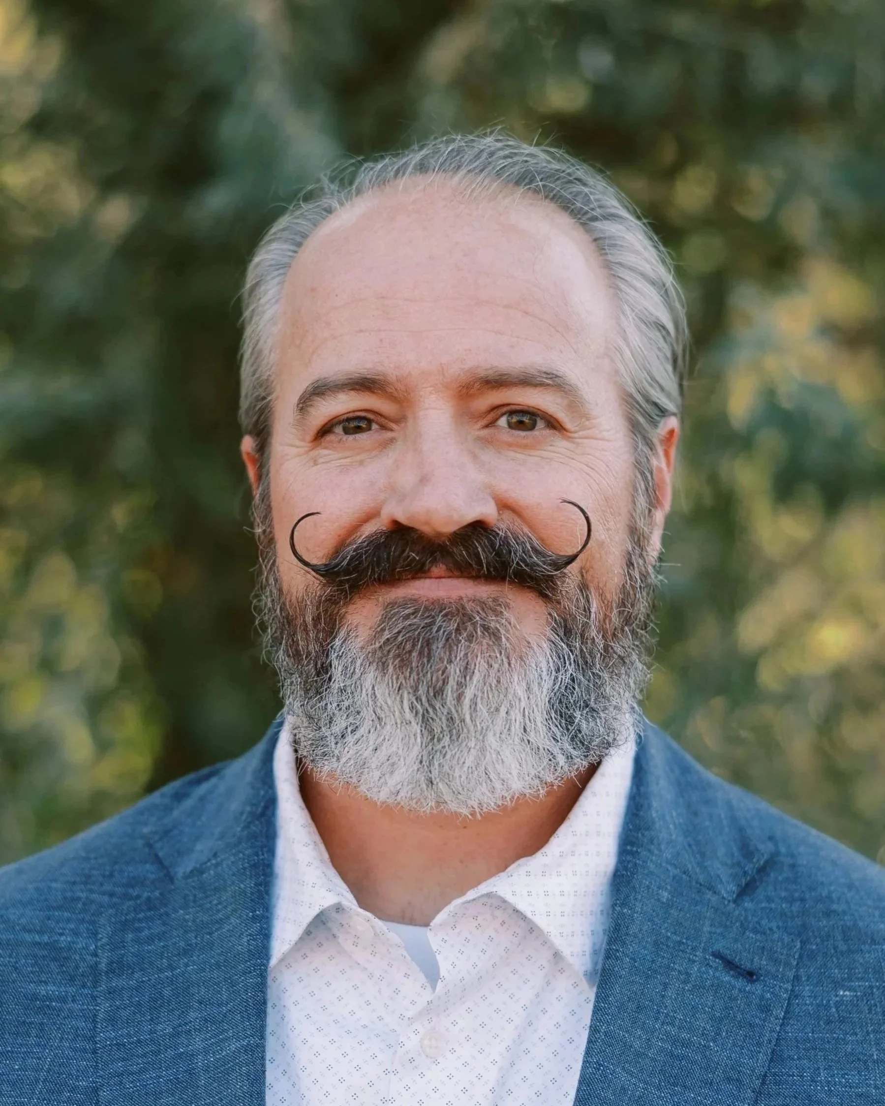 A middle-aged man with gray hair, a full beard, and styled mustache, wearing a blue blazer over a white collared shirt, standing outdoors with a blurred background of trees and foliage.