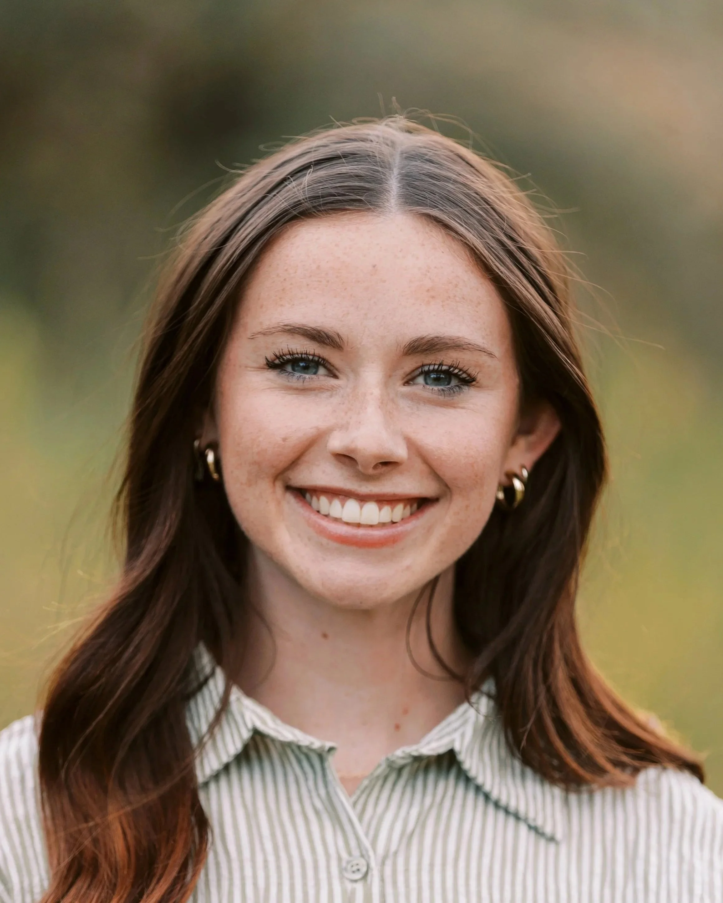 A smiling young woman with brown hair, blue eyes, and earrings, outdoors in front of a blurred green background.