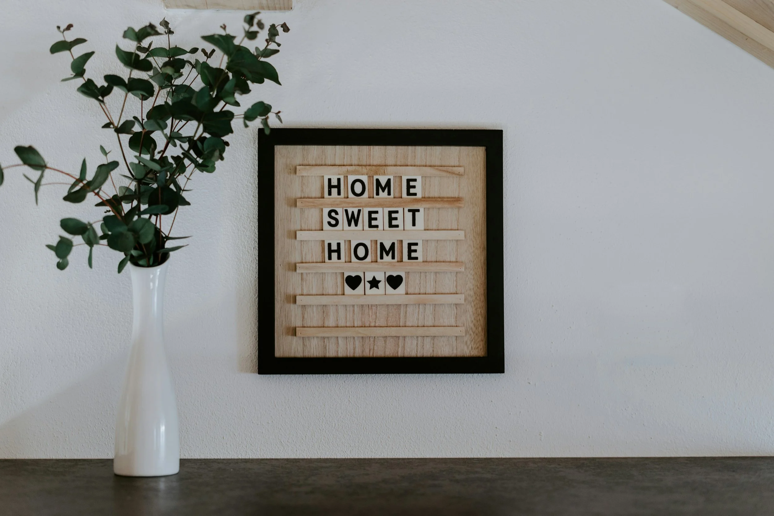 A black frame wall decor with the words 'Home Sweet Home' on horizontal wooden slats and three black icons of hearts and stars below, hanging on a white wall. To the left, a white vase with green leafy branches sits on a dark surface.