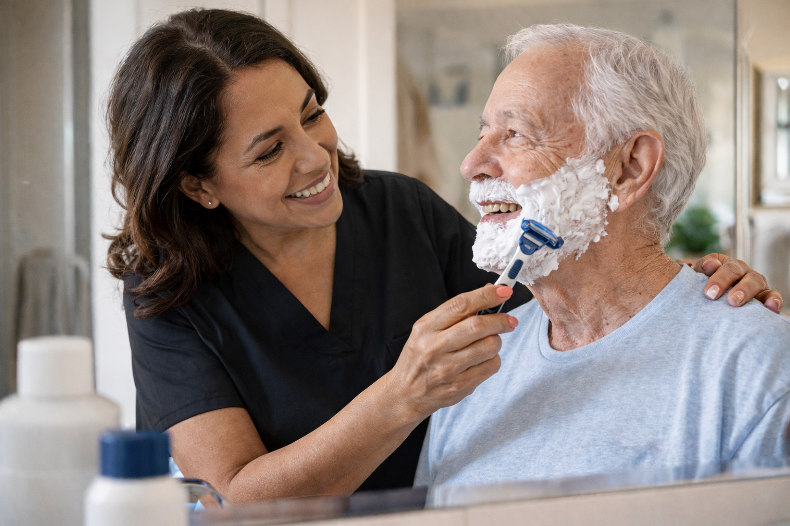 A woman assisting an elderly man with shaving, applying shaving cream on his face with a razor, both smiling at each other.