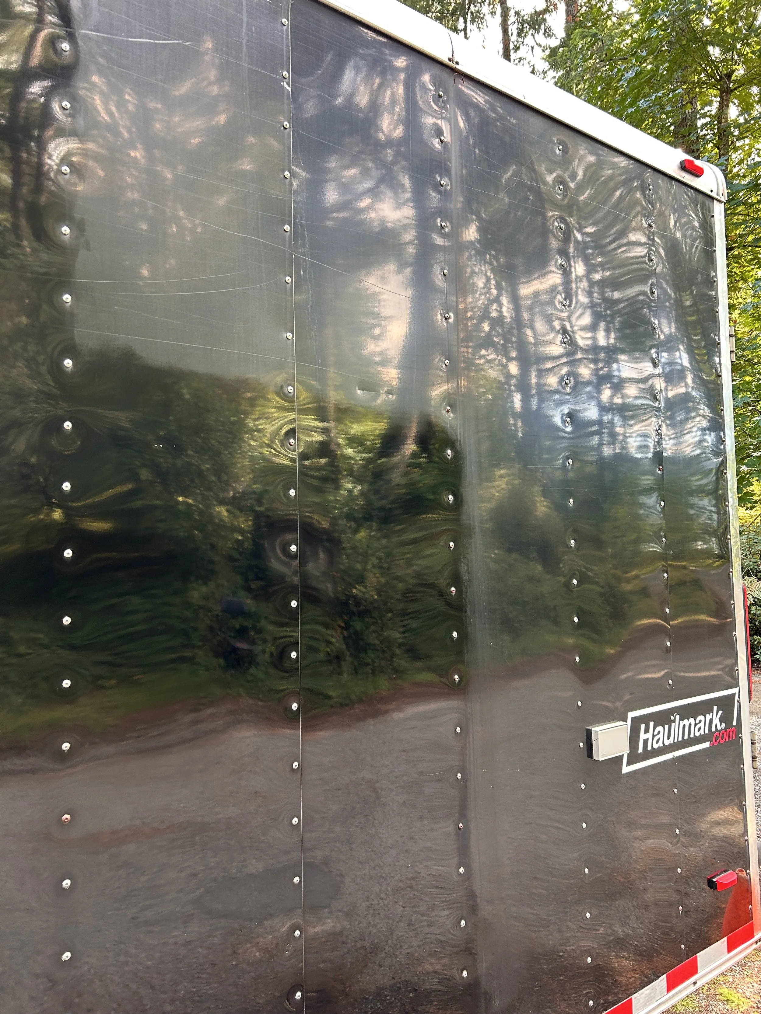 Close-up of a reflective black Haulmark trailer wall with visible rivets, trees, and sky reflected on its surface.
