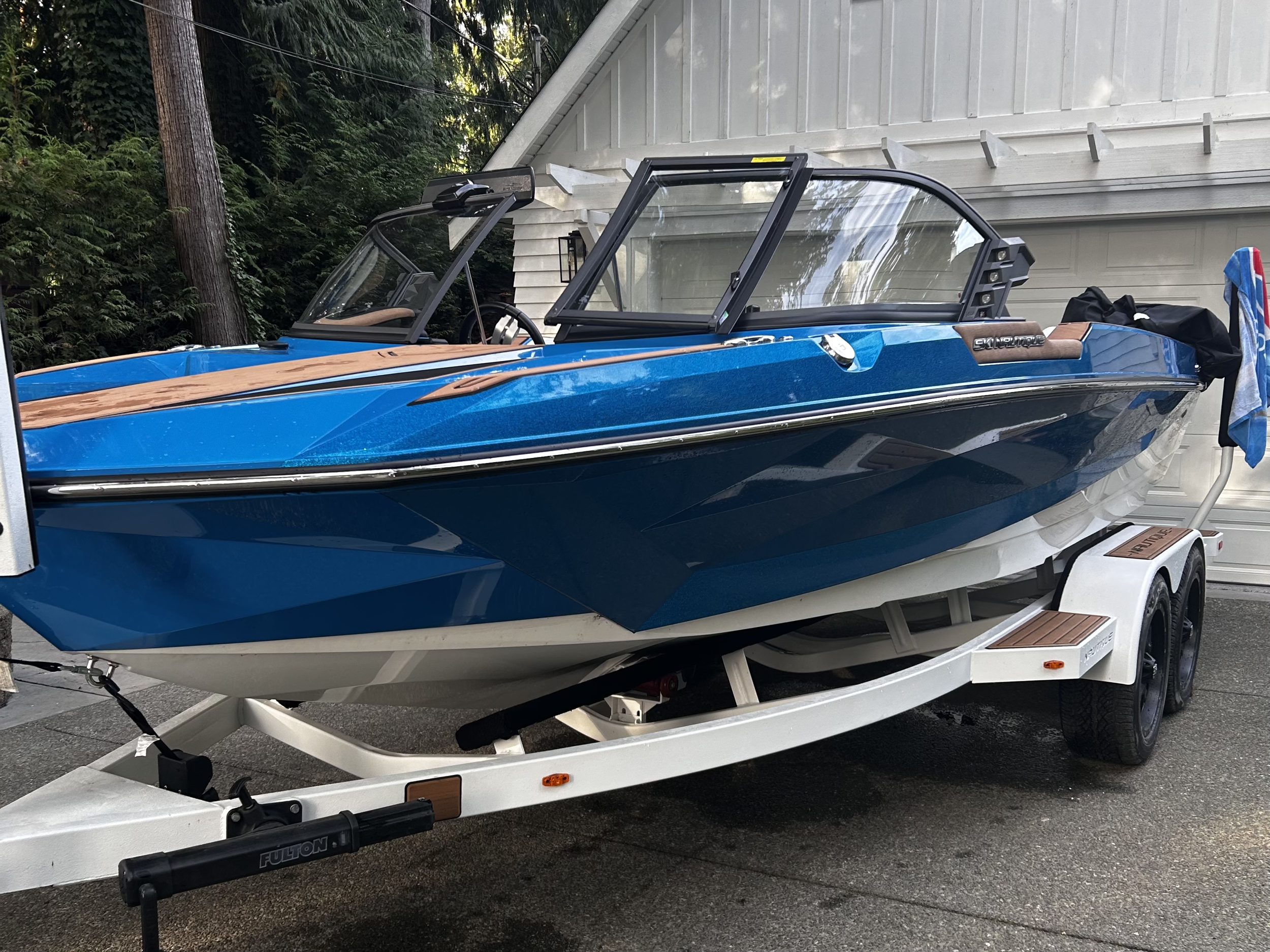 A blue and white speedboat on a trailer parked in front of a white garage door, with a towel hanging on the trailer's side.