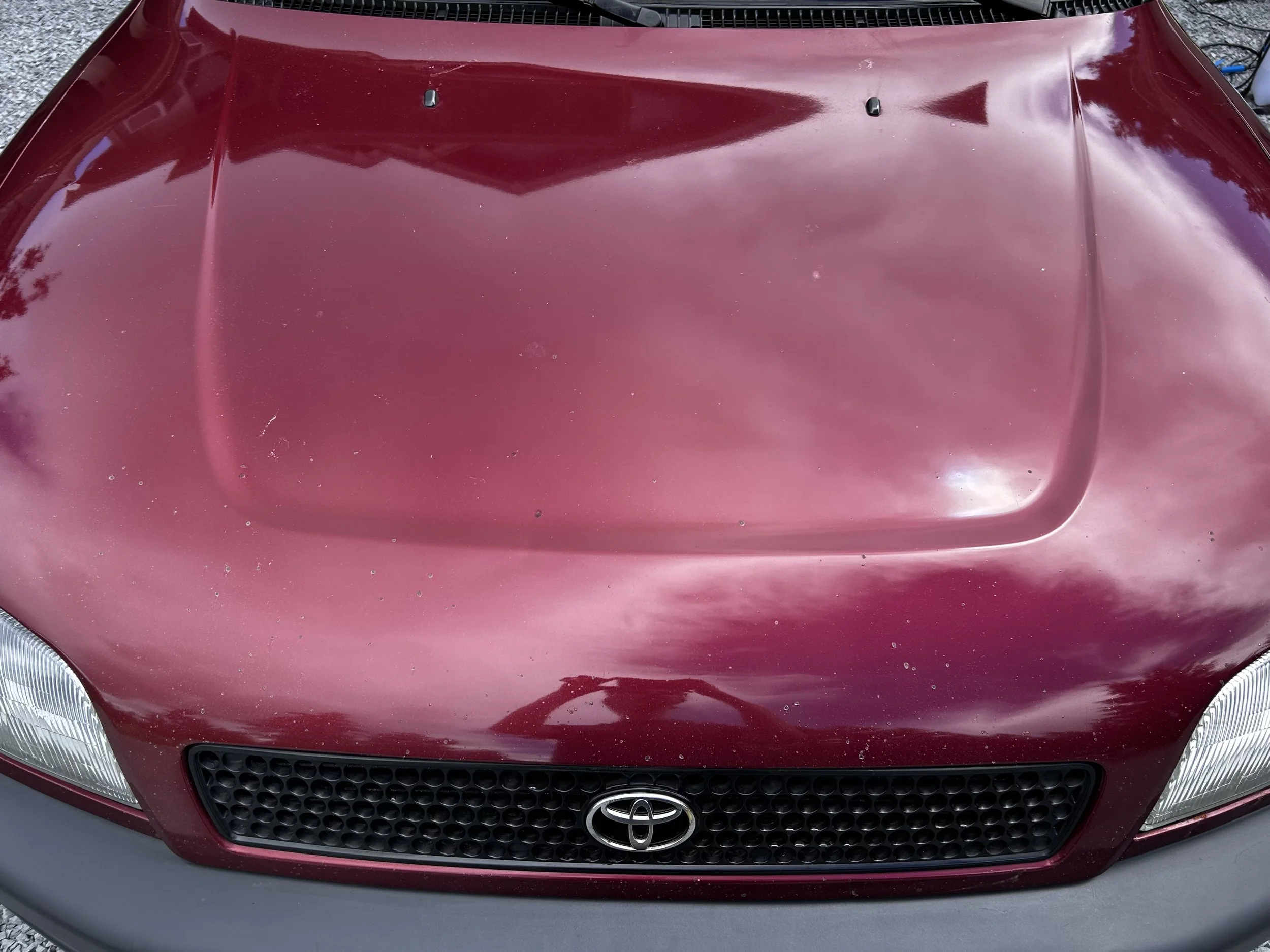 Close-up of a red Toyota car hood with a raised scoop, some dirt and debris, and reflections of the sky and clouds.