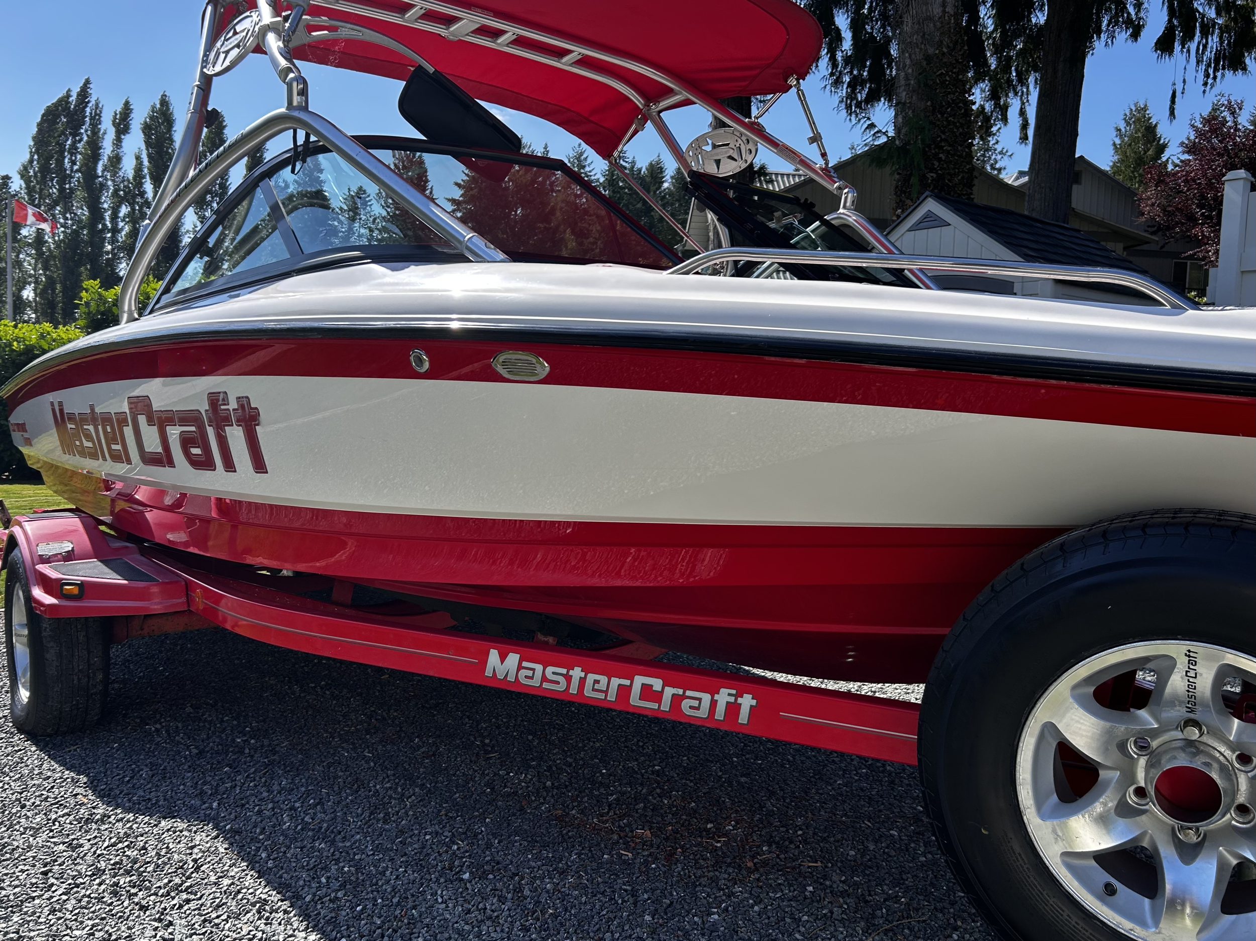 Red and white MasterCraft speedboat on a matching red trailer parked on a gravel surface, with trees and houses in the background.