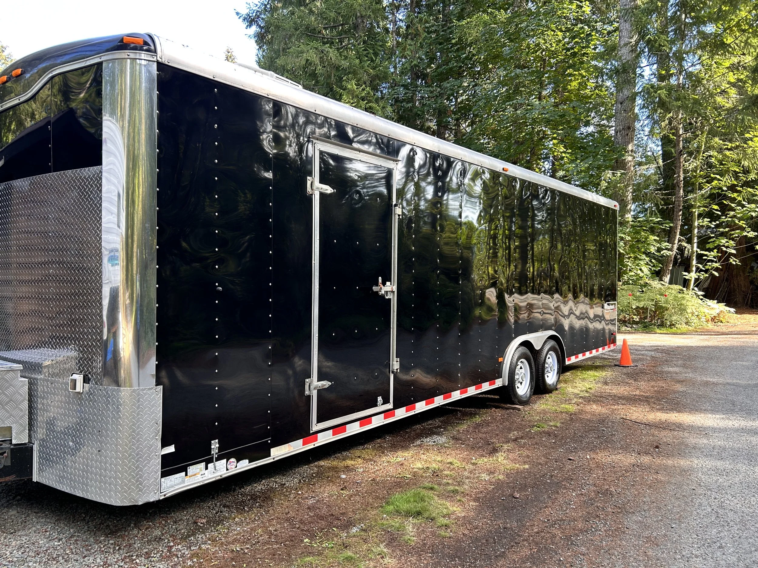 Black enclosed trailer with double axles, parked on a gravel and dirt surface, with a small orange traffic cone beside it, surrounded by green trees.