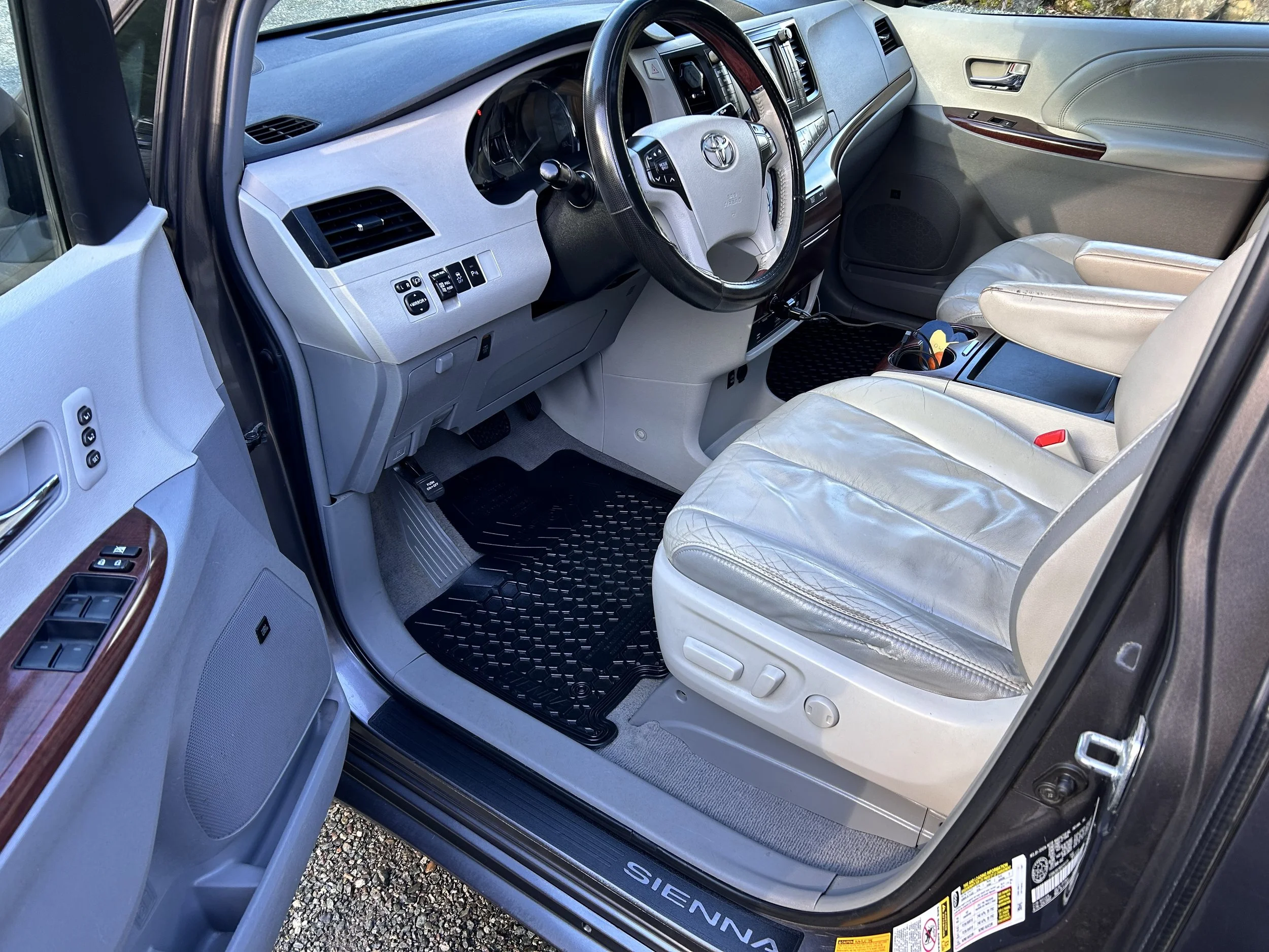 Interior of a Toyota Sienna minivan showing the driver's seat, steering wheel, dashboard, and passenger seat with some wear on the leather, and black floor mats.