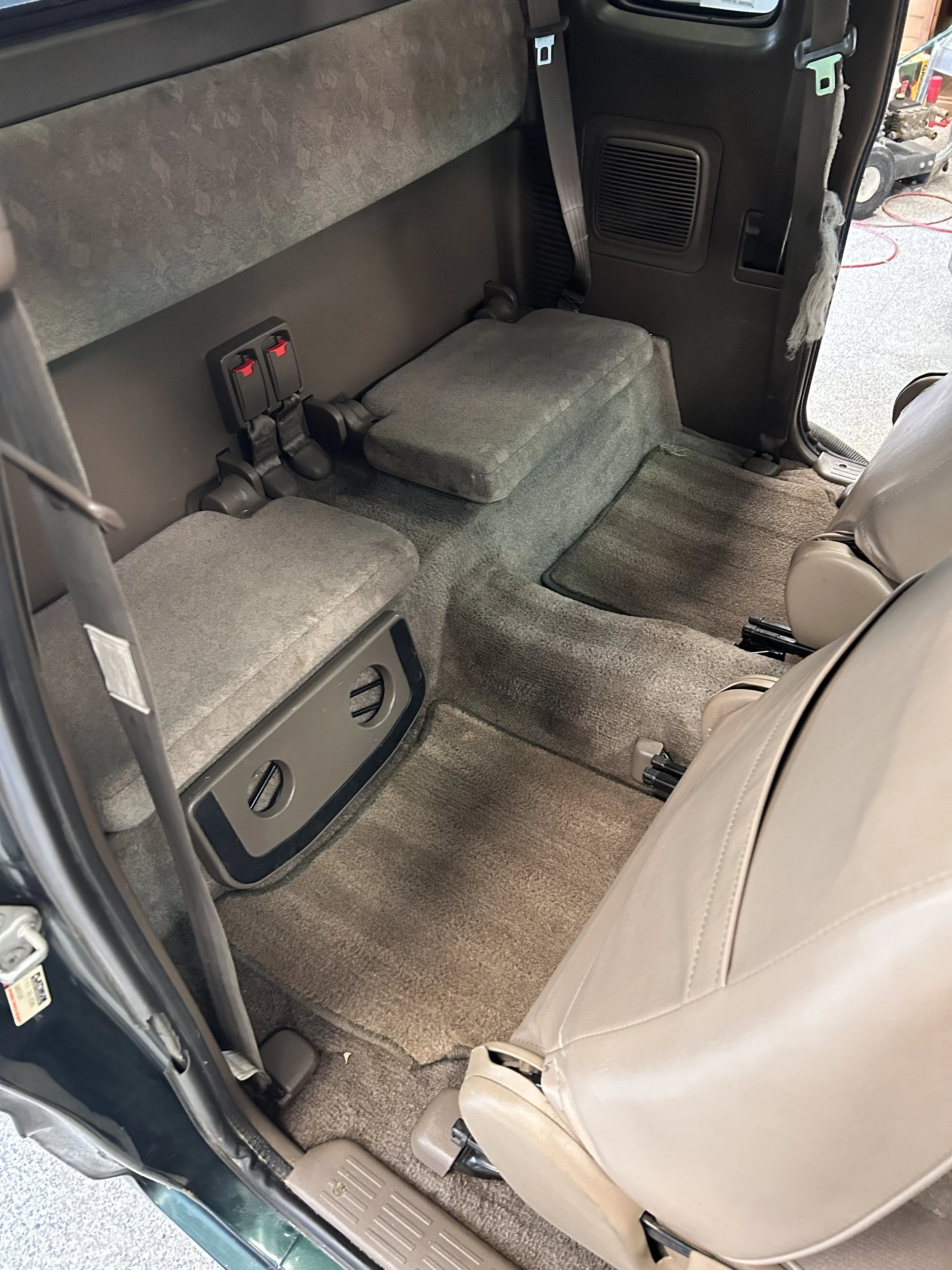 The interior of a vehicle's back seat area, with two rows of gray fabric seats, seat belts, and a beige front seat visible.