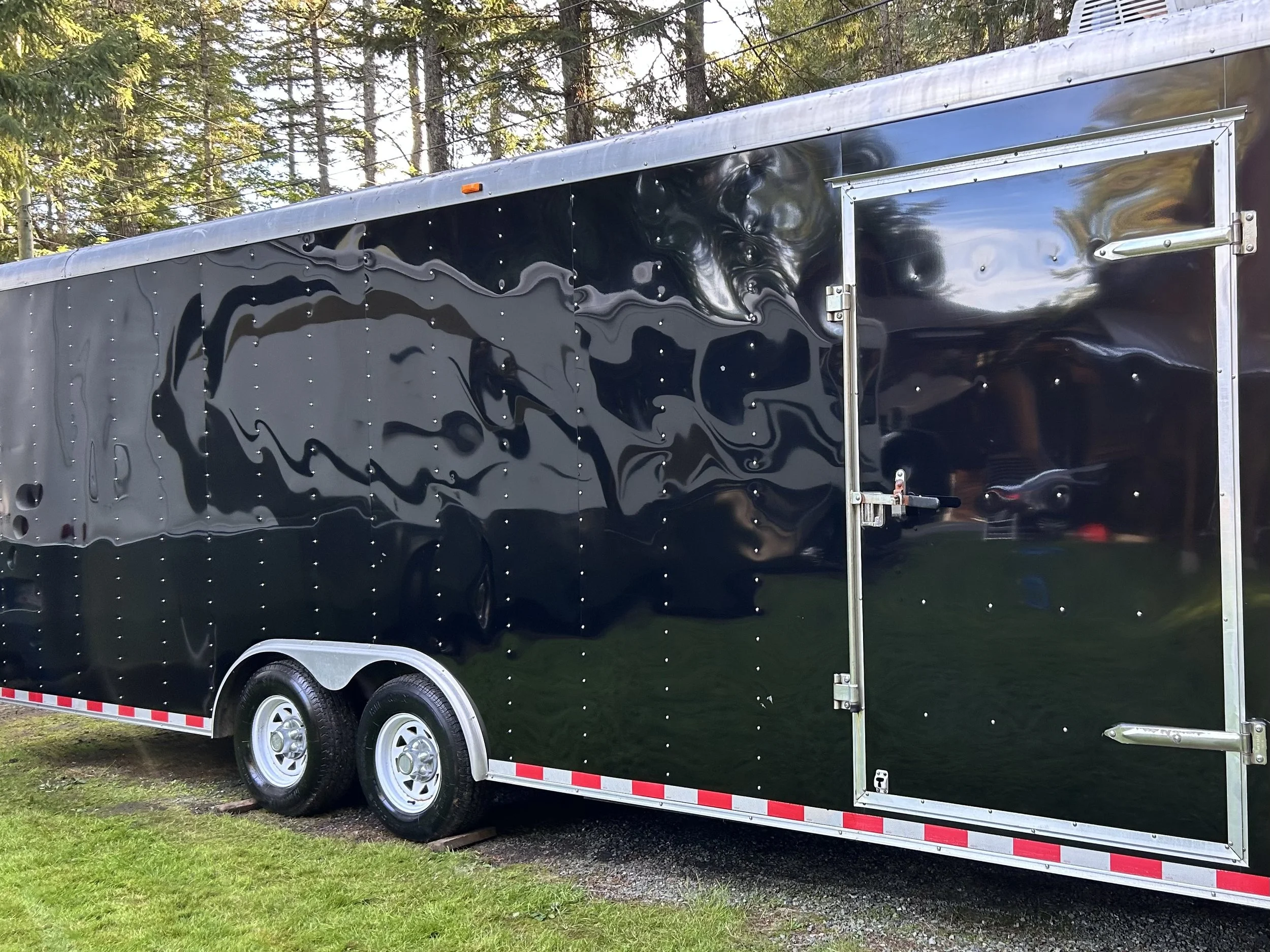 A black enclosed cargo trailer with a shiny, reflective surface, parked outdoors on grass near trees.