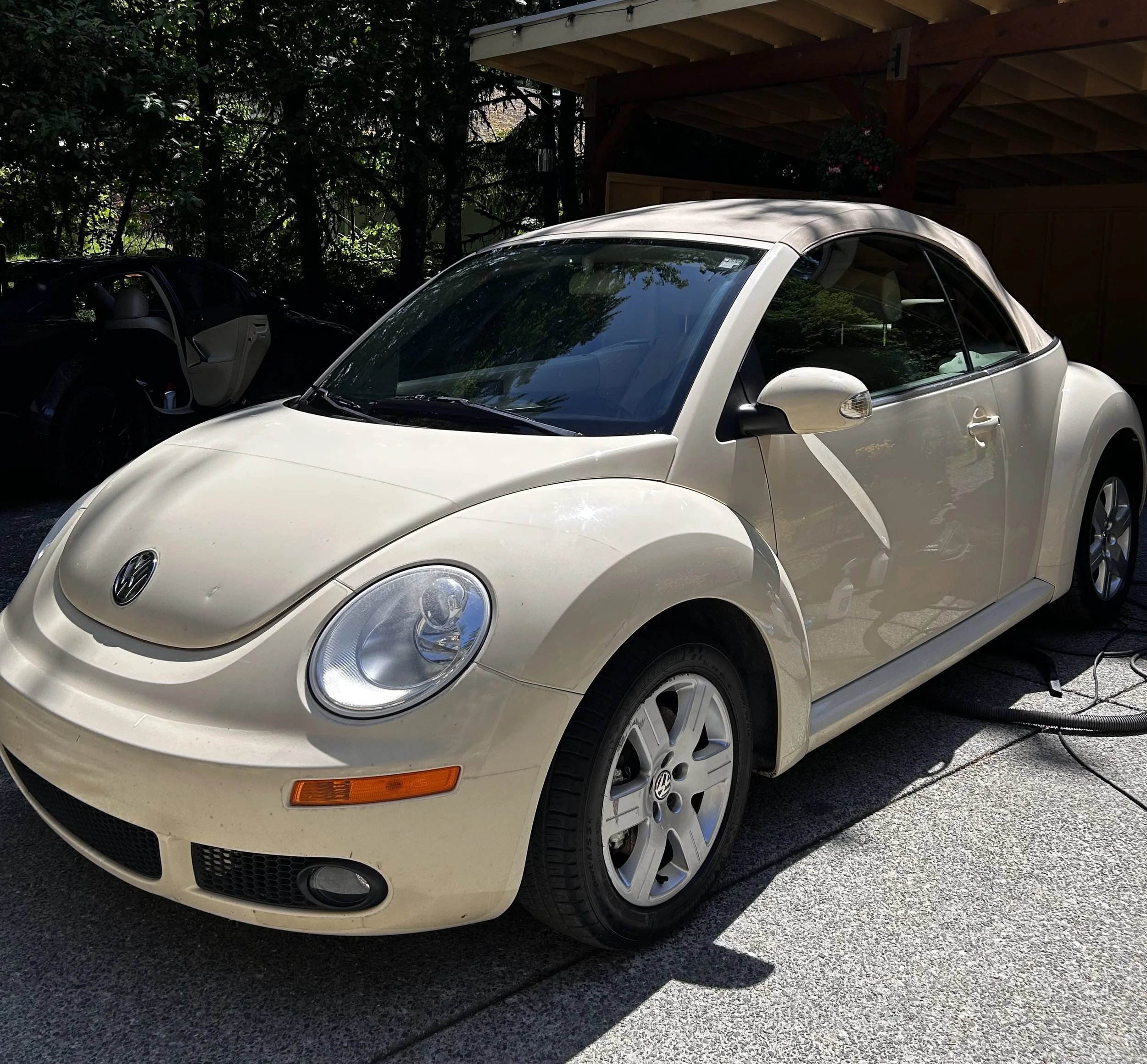 White Volkswagen Beetle convertible parked outdoors, connected to a car wash vacuum hose.