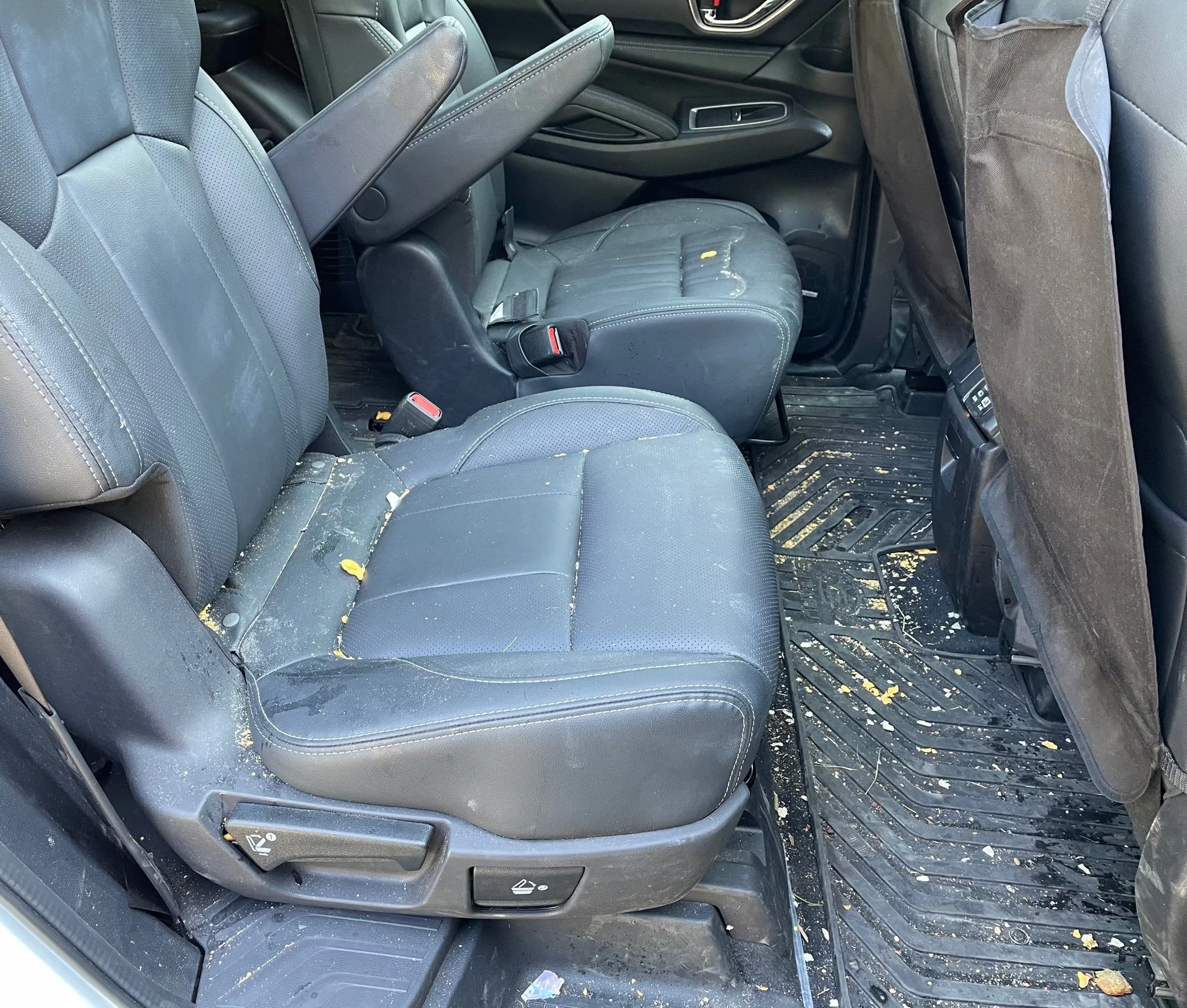 The interior of a car showing both the front and rear seats, with the seats covered in black leather or leather-like material. The seats are dirty and stained, with debris and yellowish marks scattered on the seats and floor mats.