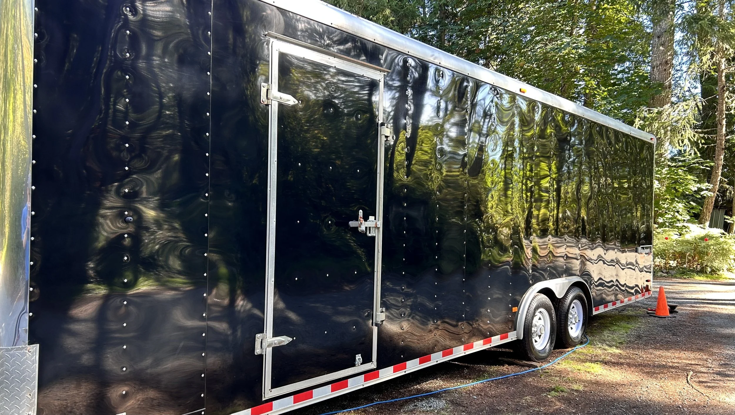 A black enclosed trailer parked outdoors among trees, with a small orange safety cone beside it.
