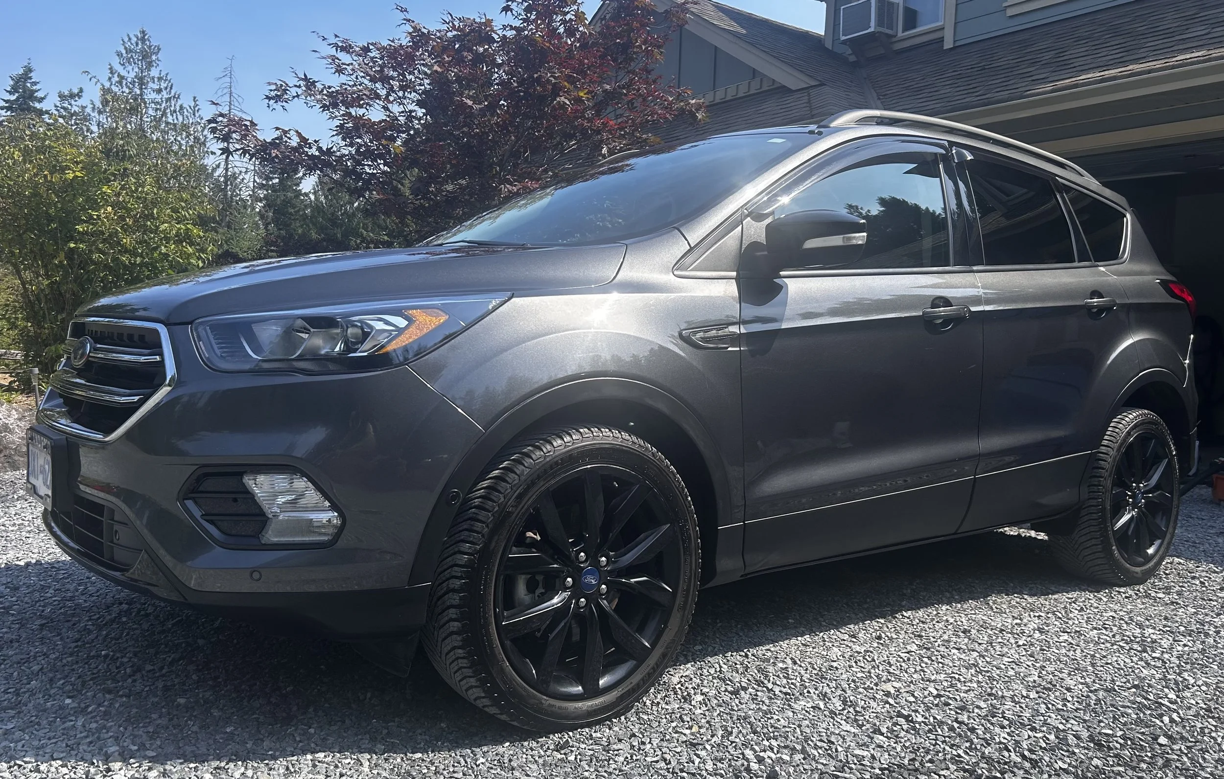 A gray Ford SUV parked on a gravel driveway with trees and a house in the background.