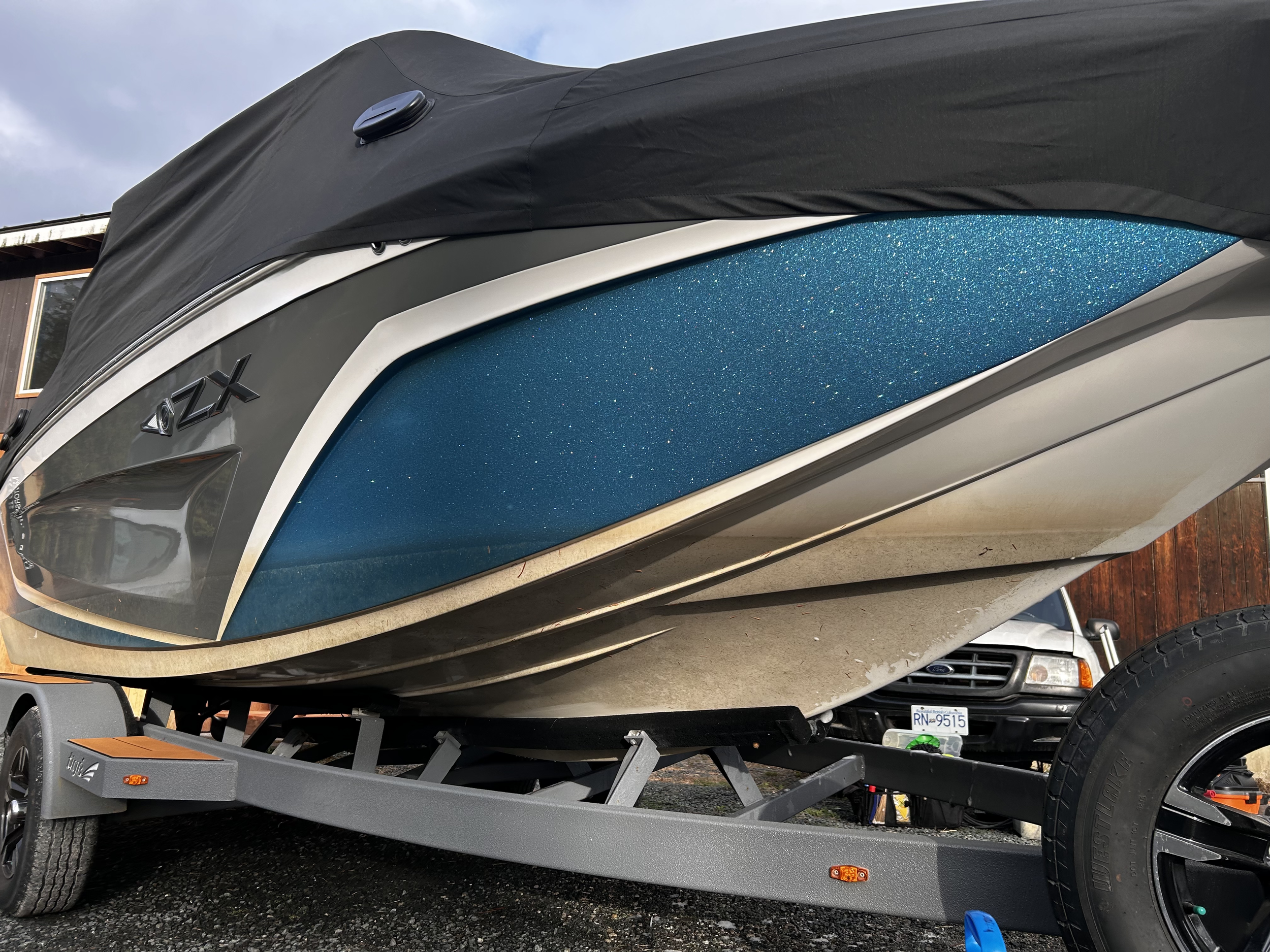 A boat on a trailer with a black cover, featuring a grey, white, and blue color scheme, parked outdoors near a wooden building with a Ford vehicle and license plate 'RN 9515' in the background.