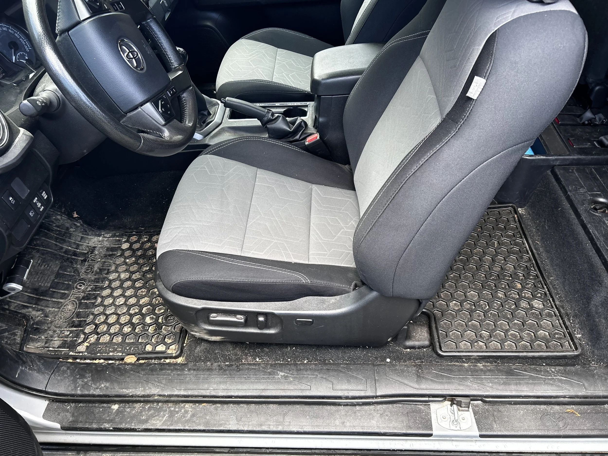 Inside view of a car showing the driver's seat, steering wheel, gear shift, and part of the front passenger's seat. The interior appears to be a Toyota vehicle with a textured rubber floor mat and dusty black interior components.