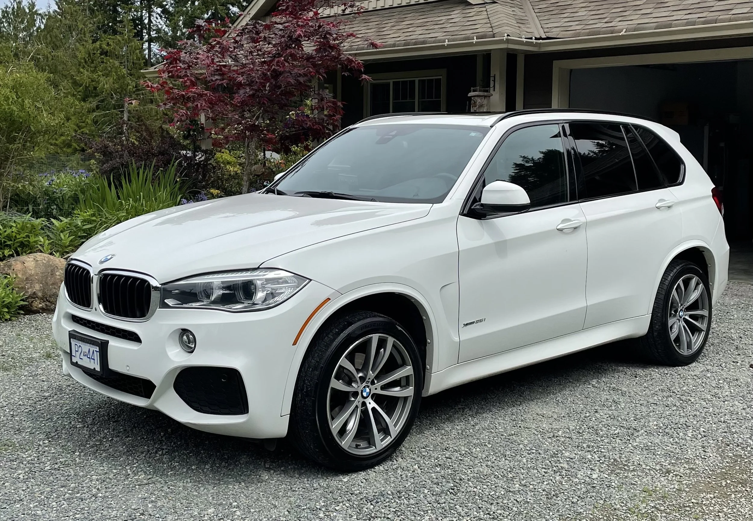 White BMW X5 SUV parked on gravel driveway in front of a house with garden and trees.