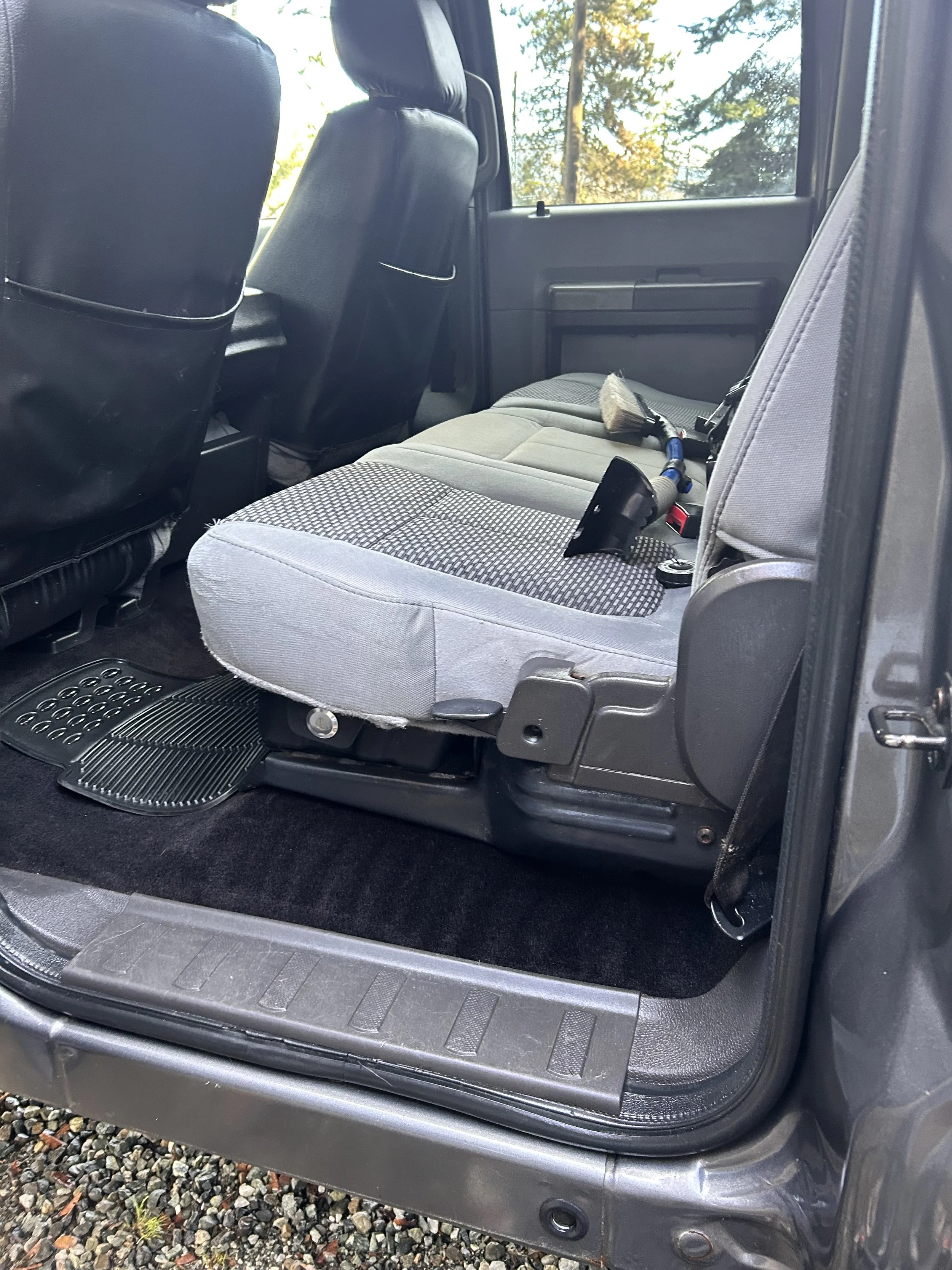 Interior of a vehicle showing a back seat with a cleaning brush, a spray bottle, and a scraper on it, with two front seats and trees visible through the window.