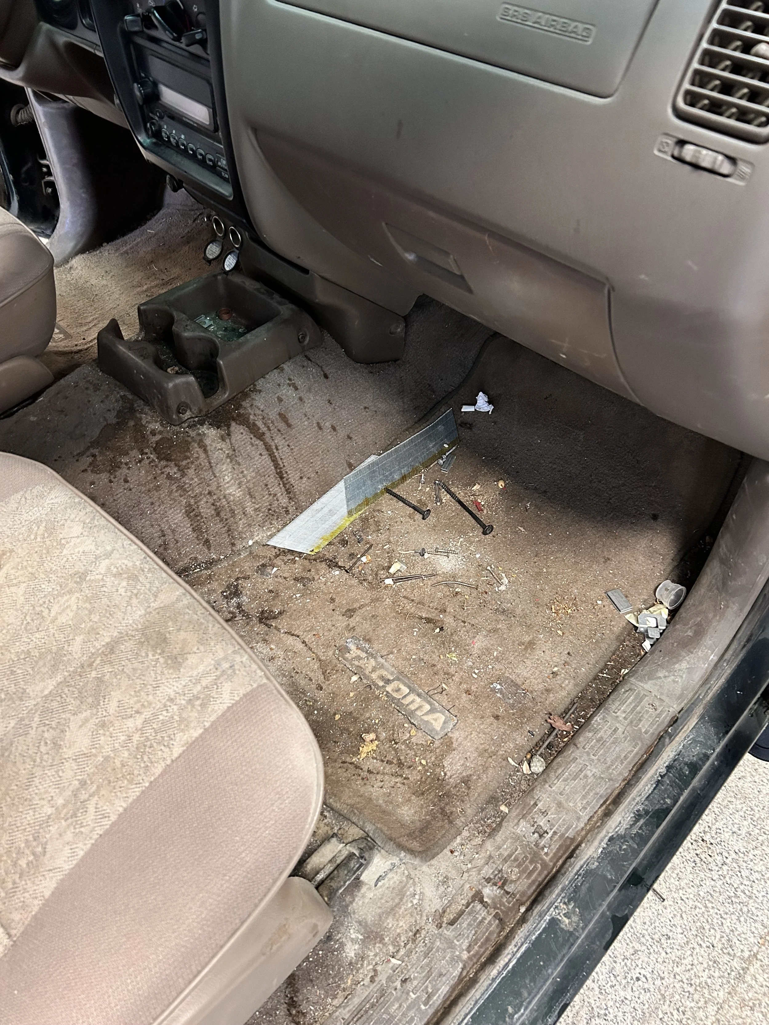 Dirty car interior with a cluttered floor, scattered trash, and tools near the driver's seat and dashboard.