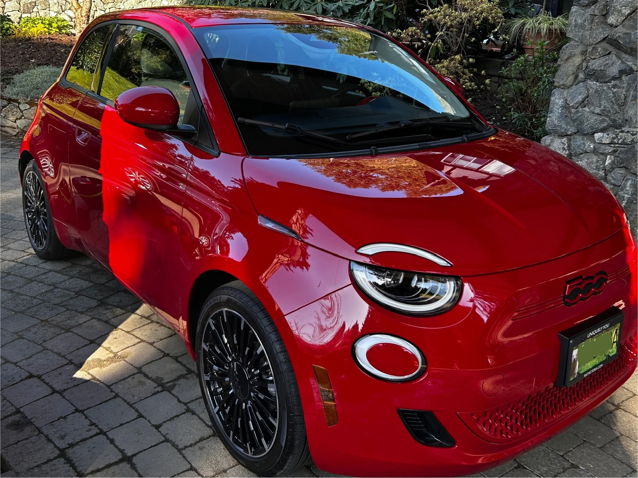 Red Fiat 500 parked on a cobblestone driveway with trees and rocks in the background.