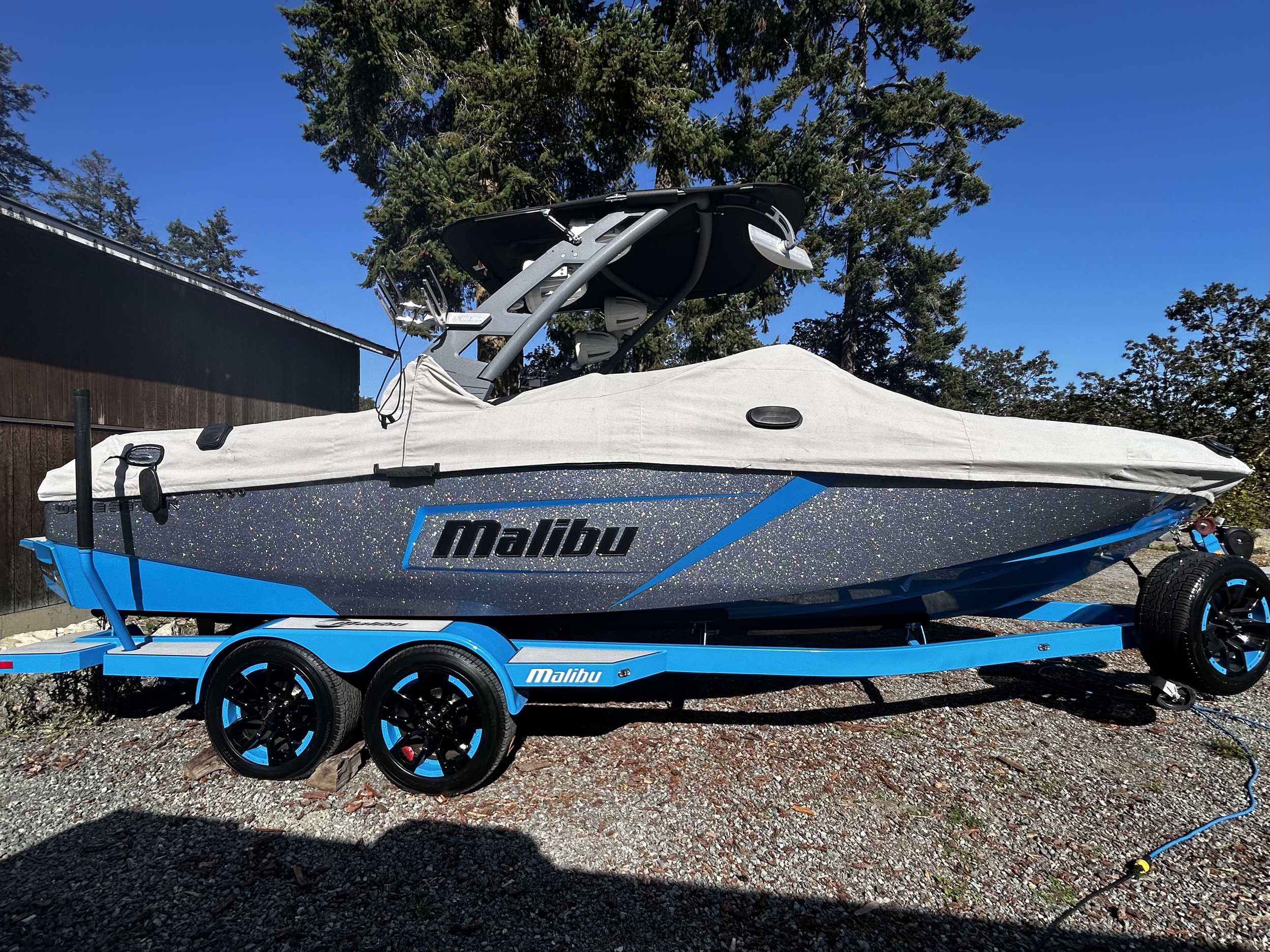A Malibu brand speedboat on a blue trailer, covered with a white cover, parked outdoors on gravel with trees and a building in the background.