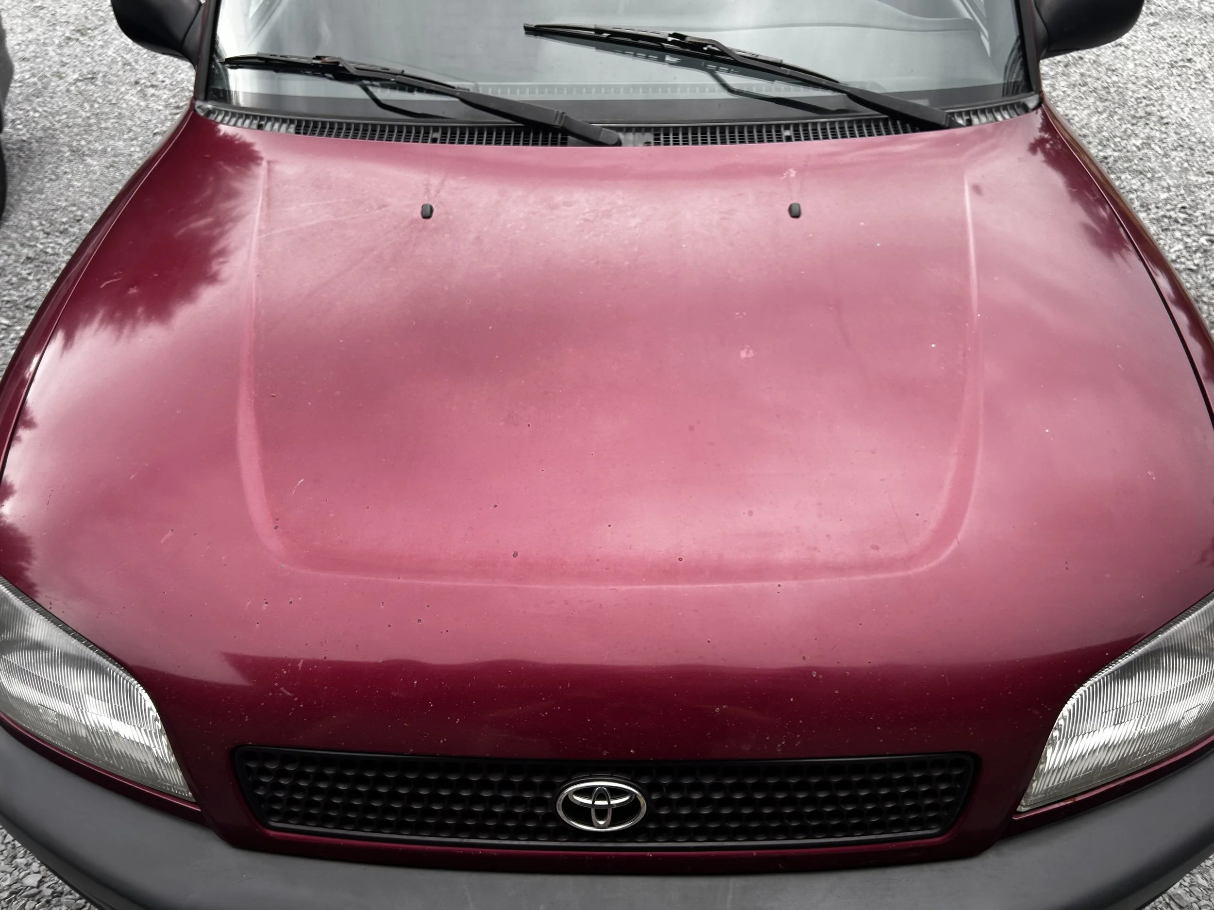 Front view of a red Toyota car with dust and dirt on the hood, parked on a gravel surface.