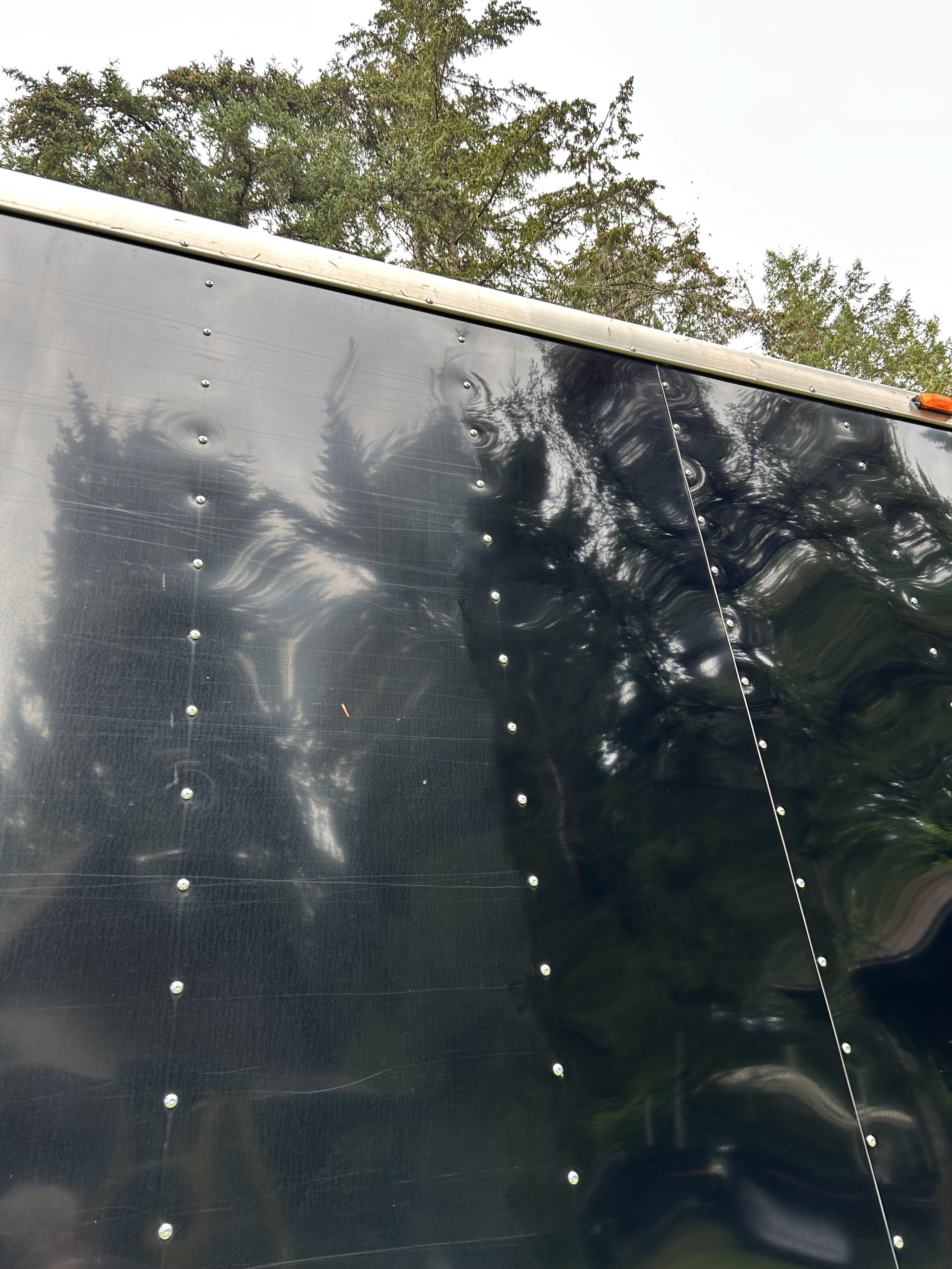 Close-up of a reflective black surface with evenly spaced bolts and ripples, with trees and sky reflected on it.
