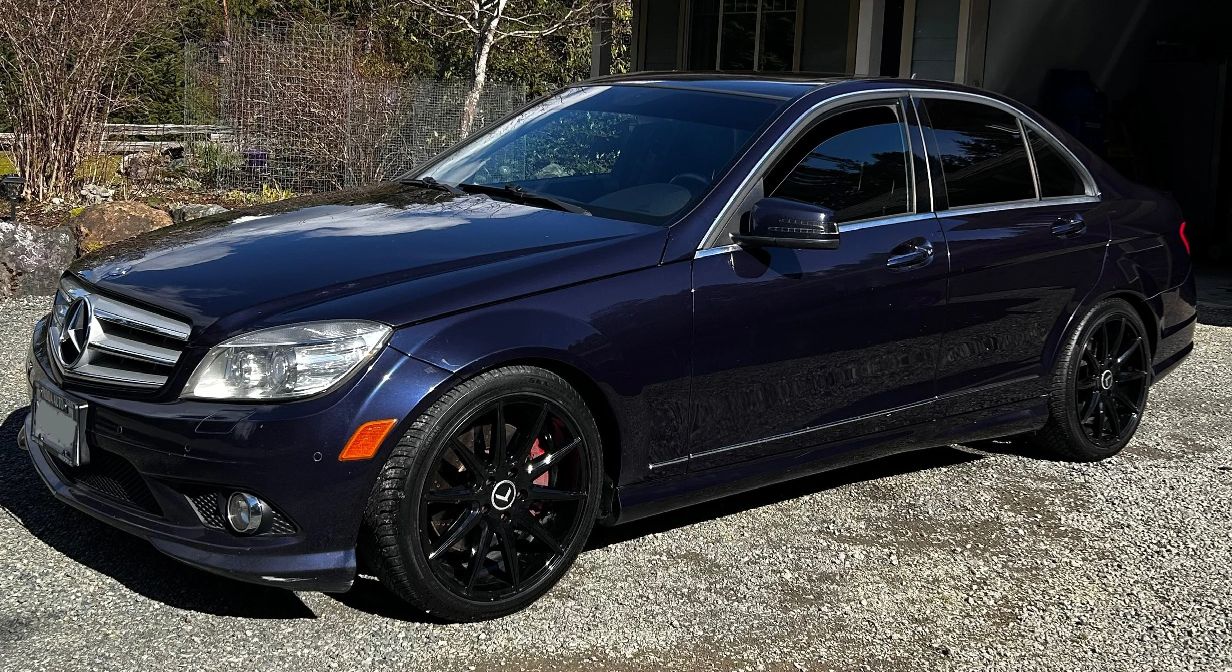 A dark blue Mercedes-Benz sedan parked on a gravel driveway with plant life and rocks in the background.