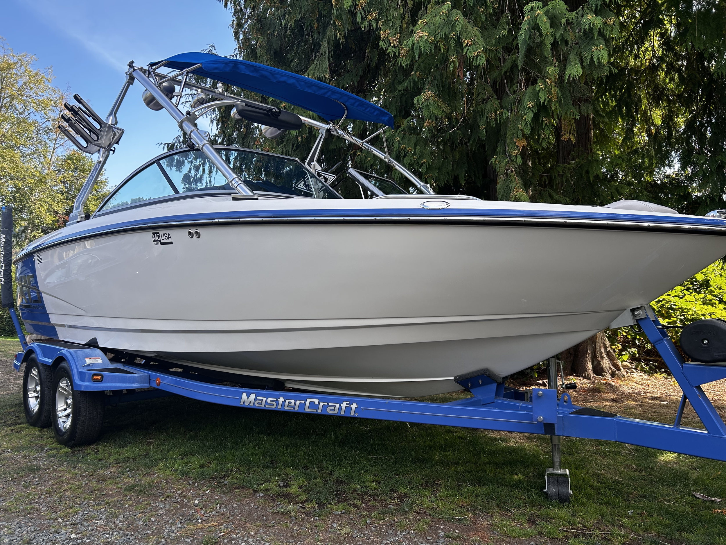 A white and blue speedboat on a blue MasterCraft trailer parked on grass with trees background.