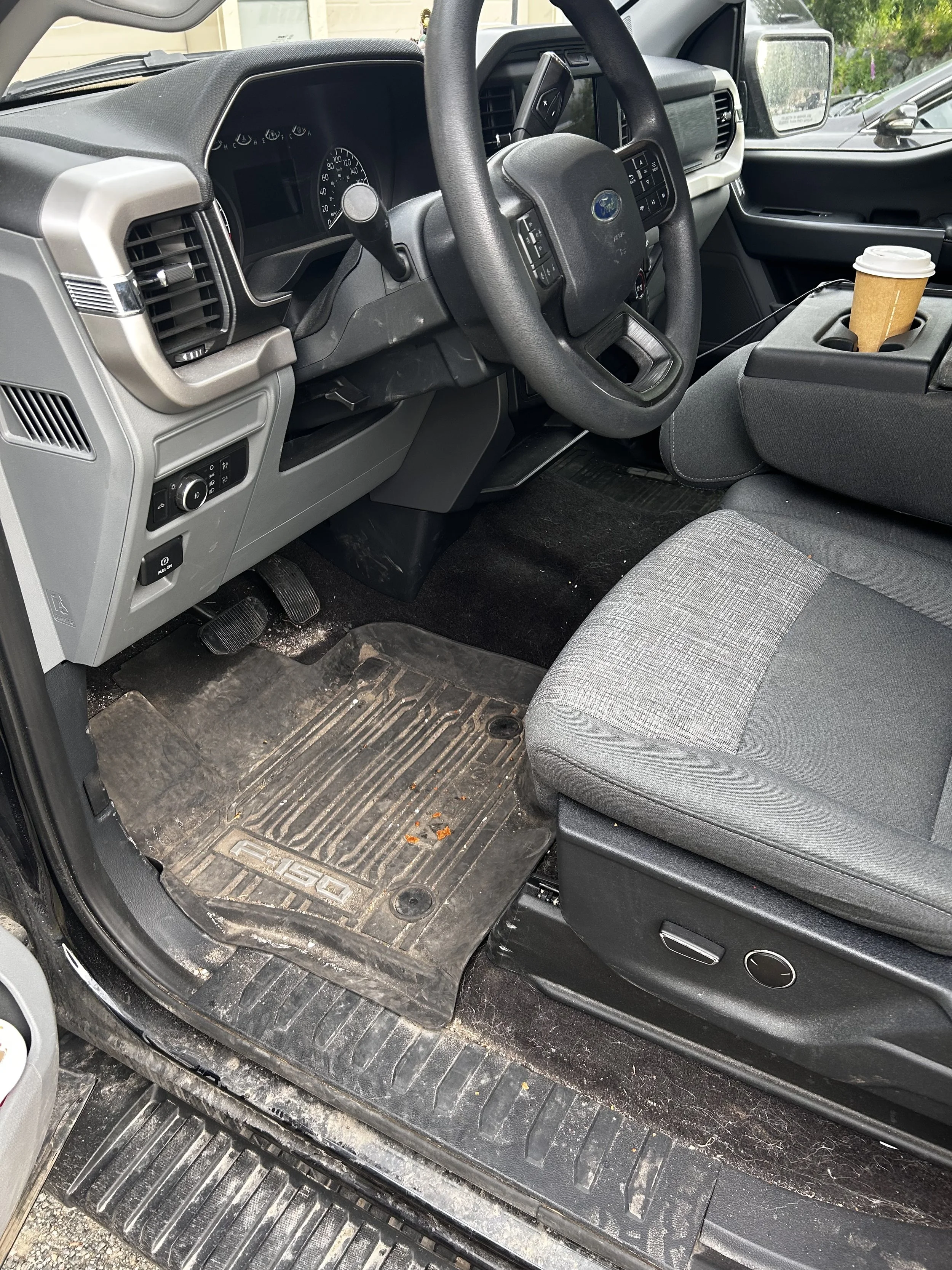 The interior of a vehicle with a gray dashboard, black steering wheel, gray seat fabric, and a coffee cup in the center console cup holder. The floor is dirty with a rubber mat that has the brand 'F-150' on it.