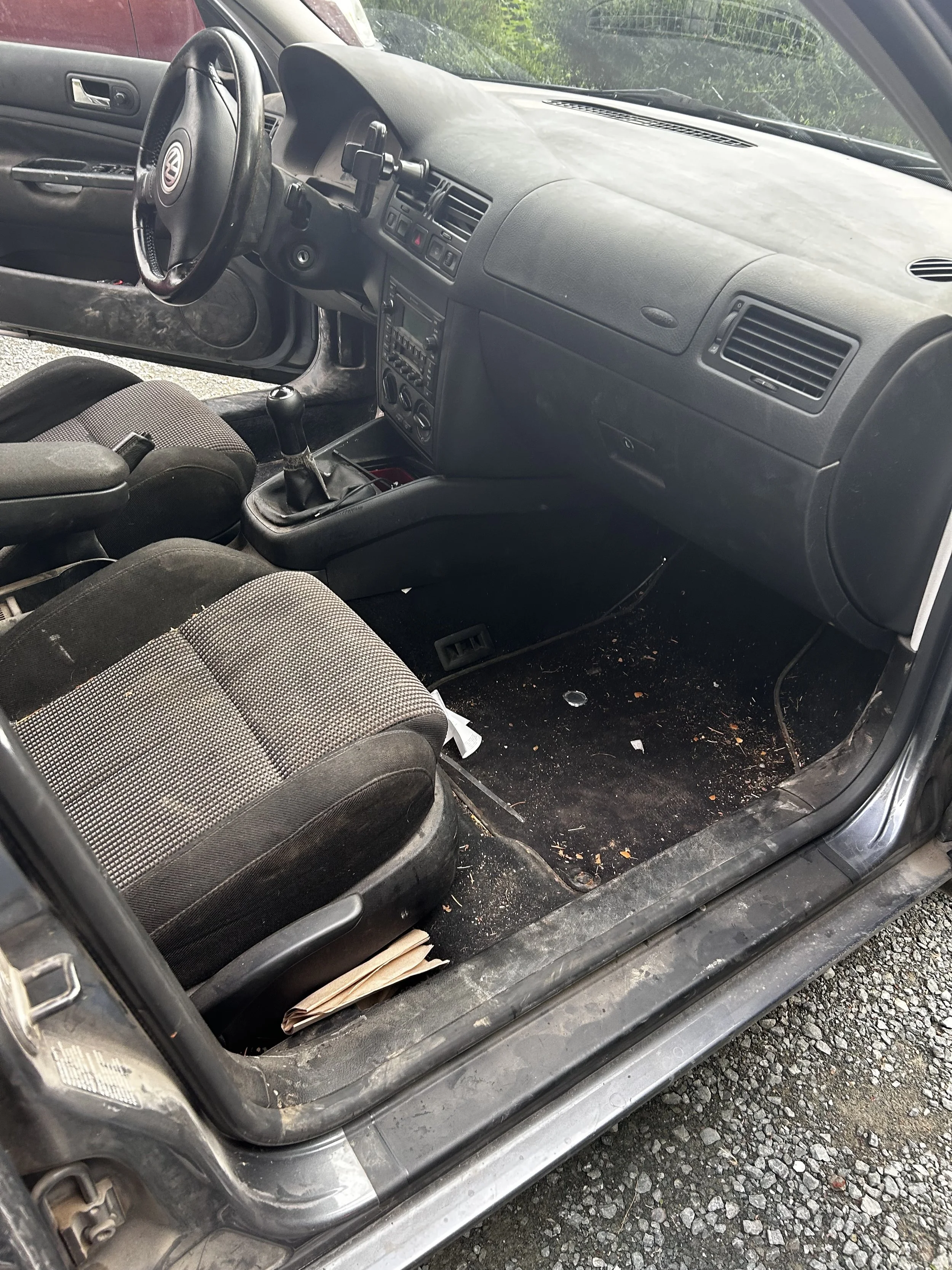 Inside a Volkswagen car showing a black dashboard, steering wheel, gear shift, two front seats with fabric upholstery, and a dirty floor with scattered debris and a folded paper underneath the front seat.