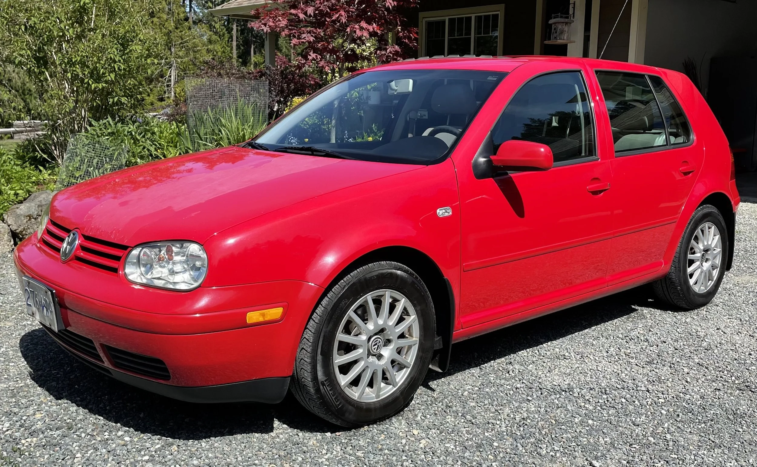 Red Volkswagen Golf parked on a gravel driveway with greenery and a house in the background.