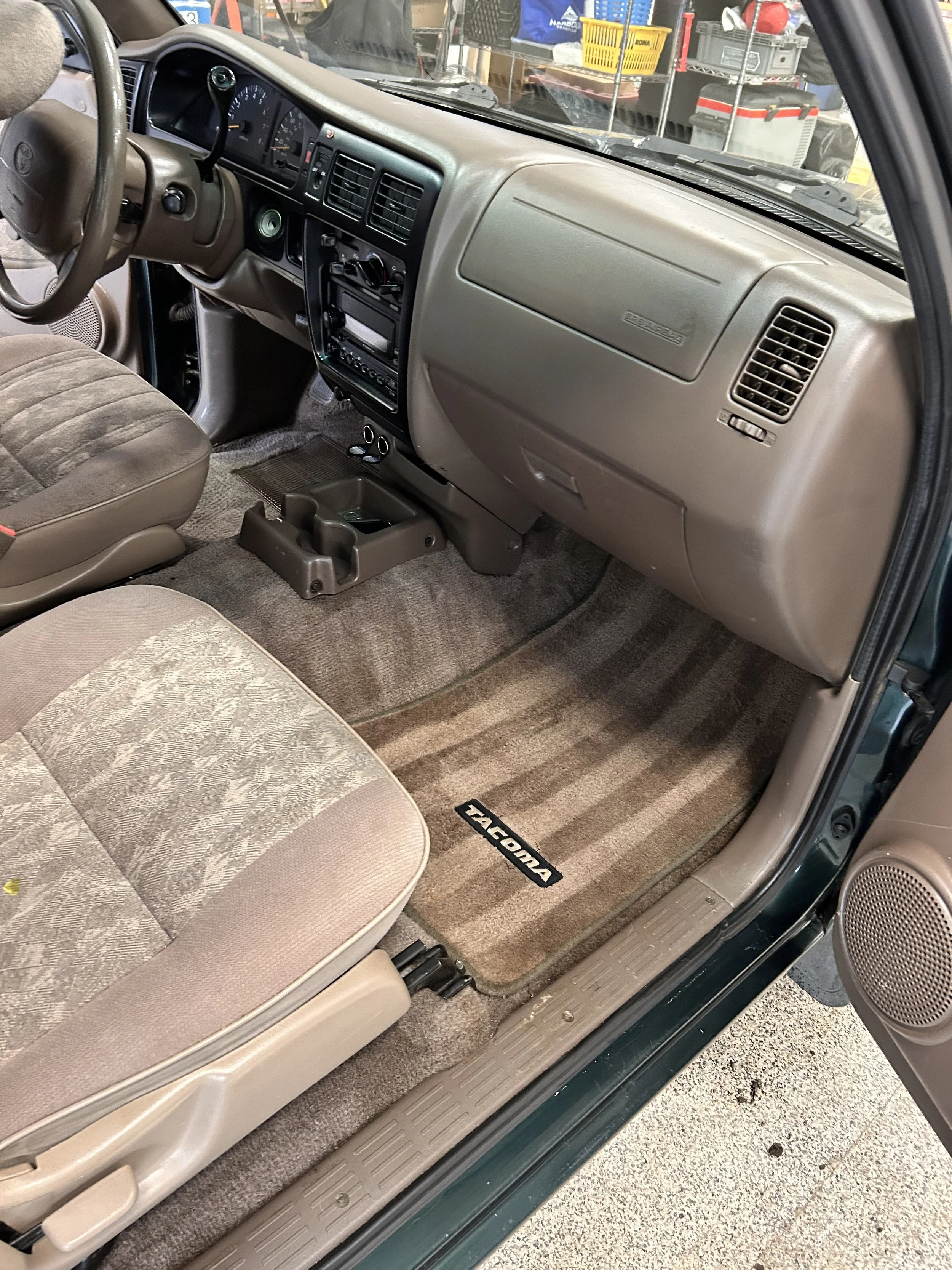 Interior of a Toyota Tacoma truck, featuring beige dashboard, steering wheel, fabric seat, center console with cup holders, and a beige floor mat with the Tacoma logo.