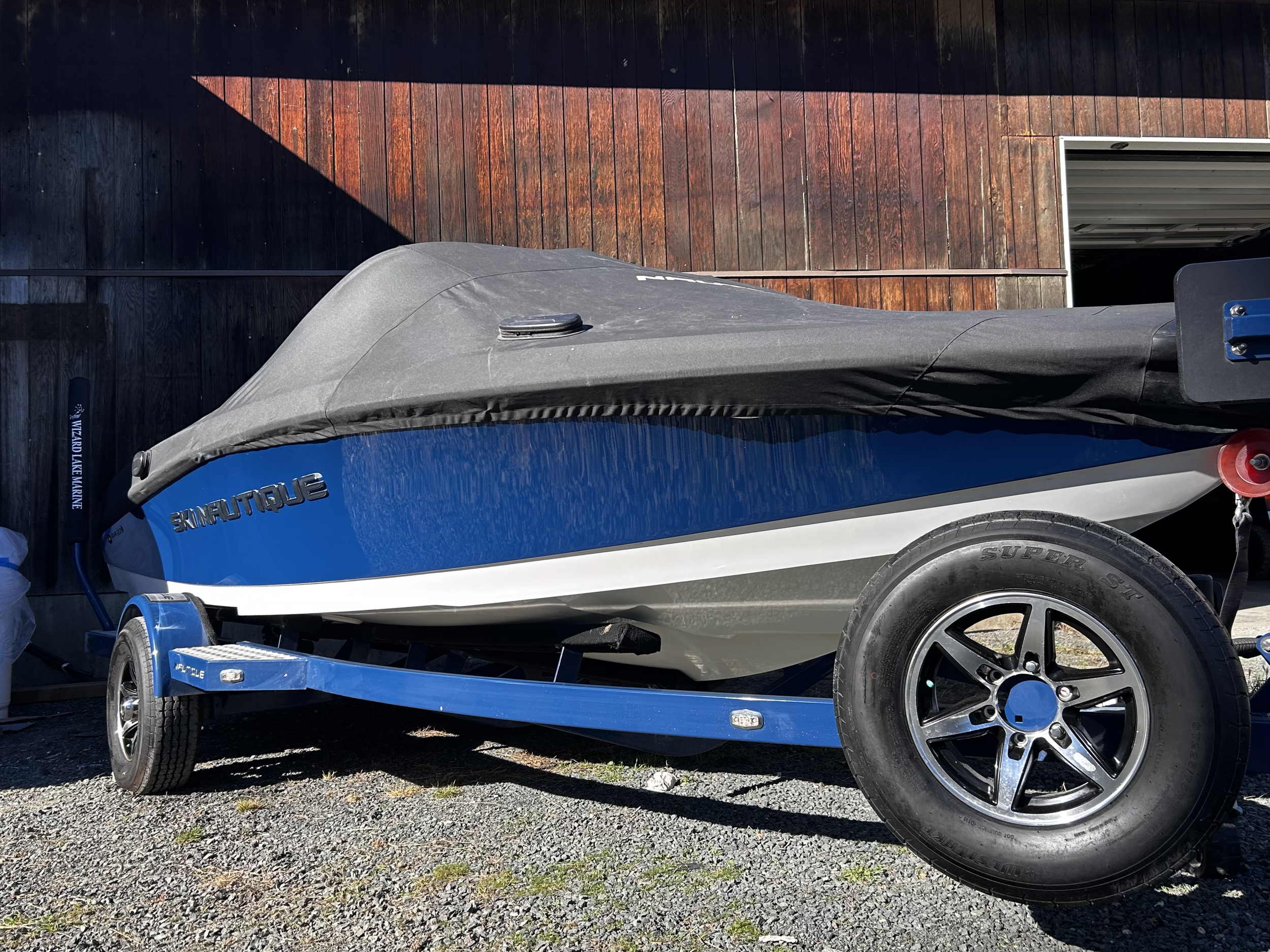 A small blue and gray boat on a blue trailer, covered with a black protective cover, parked on gravel ground in front of a wooden barn.
