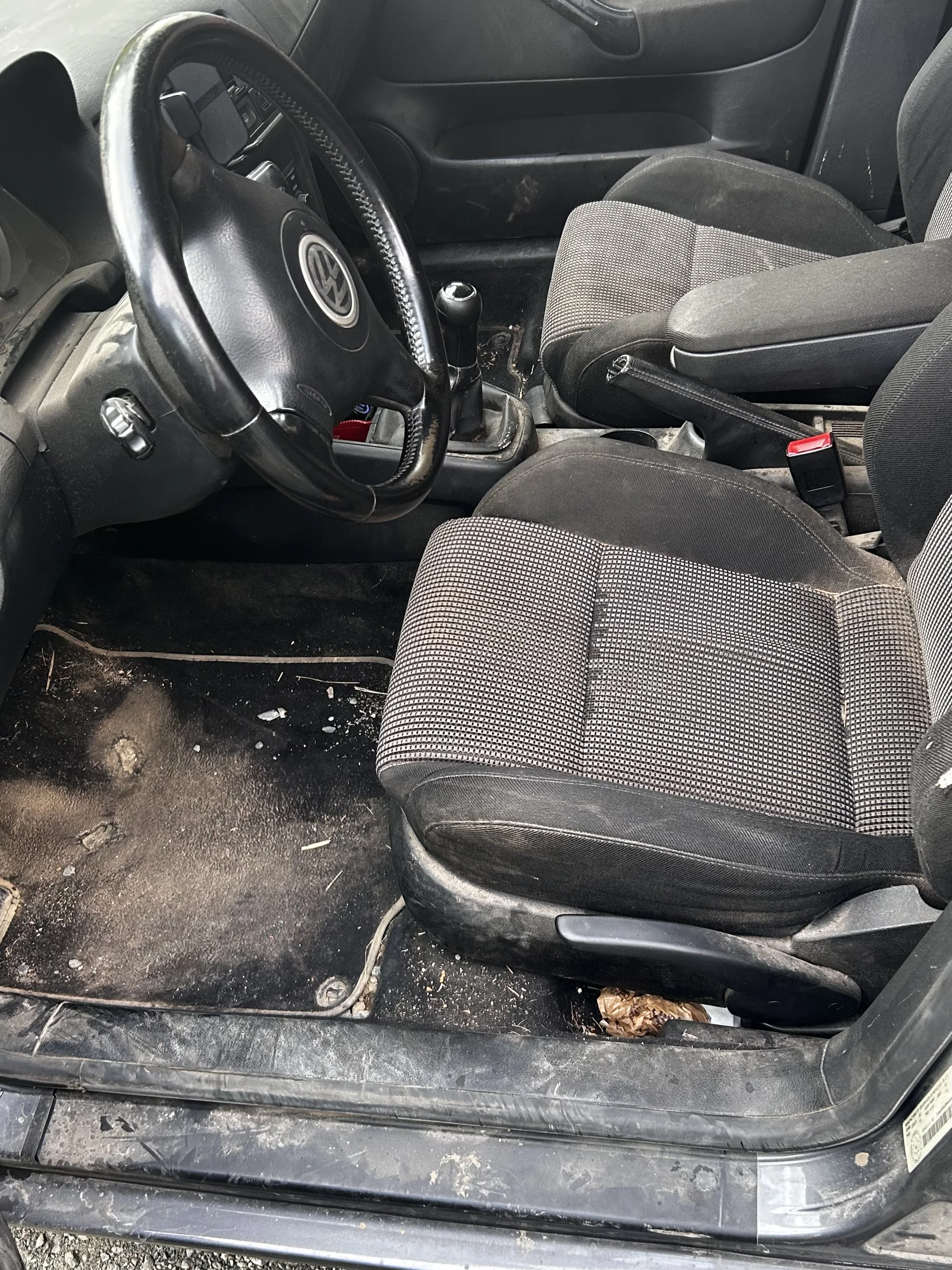 Interior of a car with a dirty black floor and seats, a manual gear shift, and a Volkswagen logo on the steering wheel.