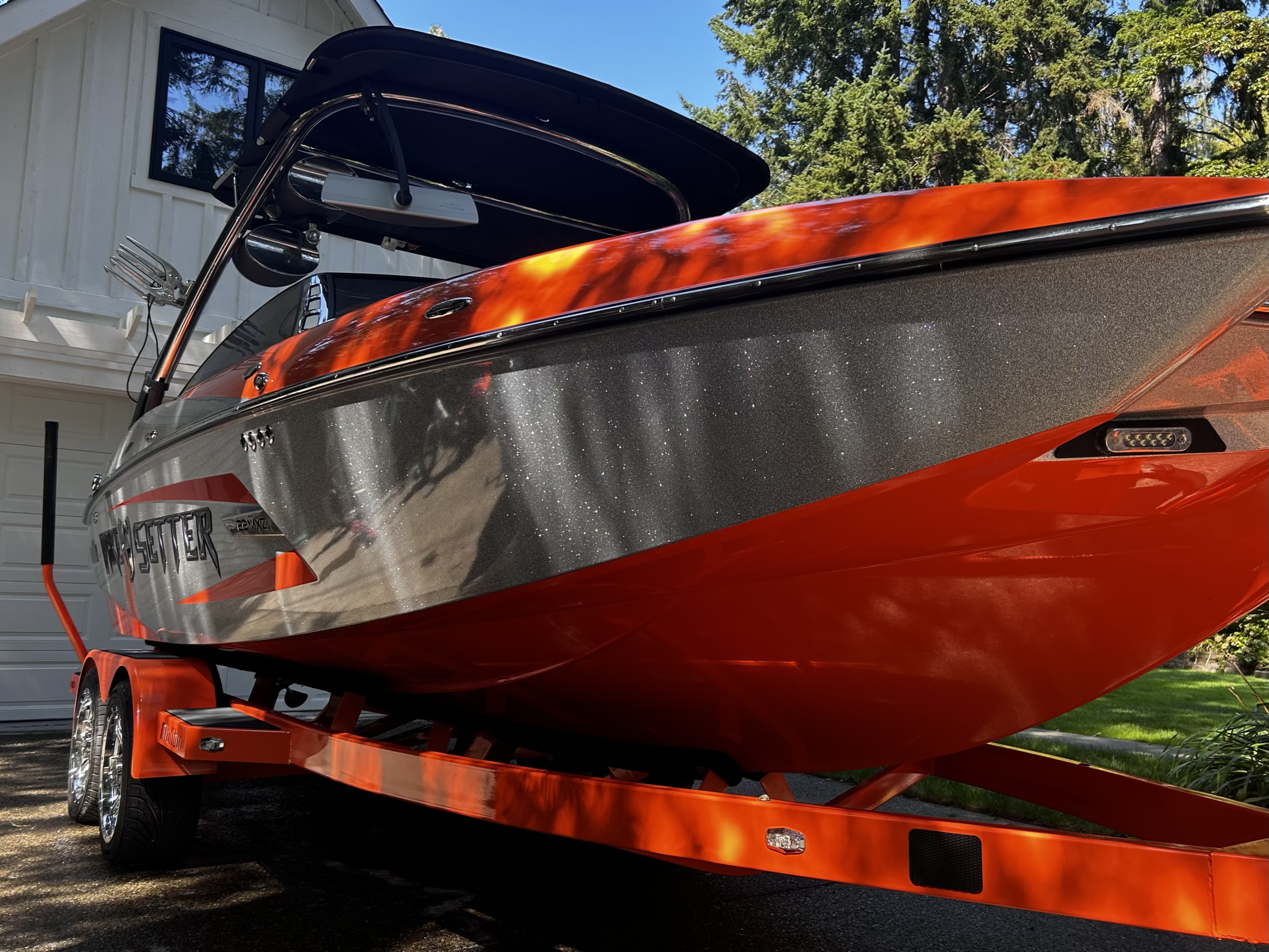 A speedboat with a shiny metallic body and orange accents, mounted on a matching orange trailer parked outside a house with white walls and black windows, surrounded by trees and greenery.