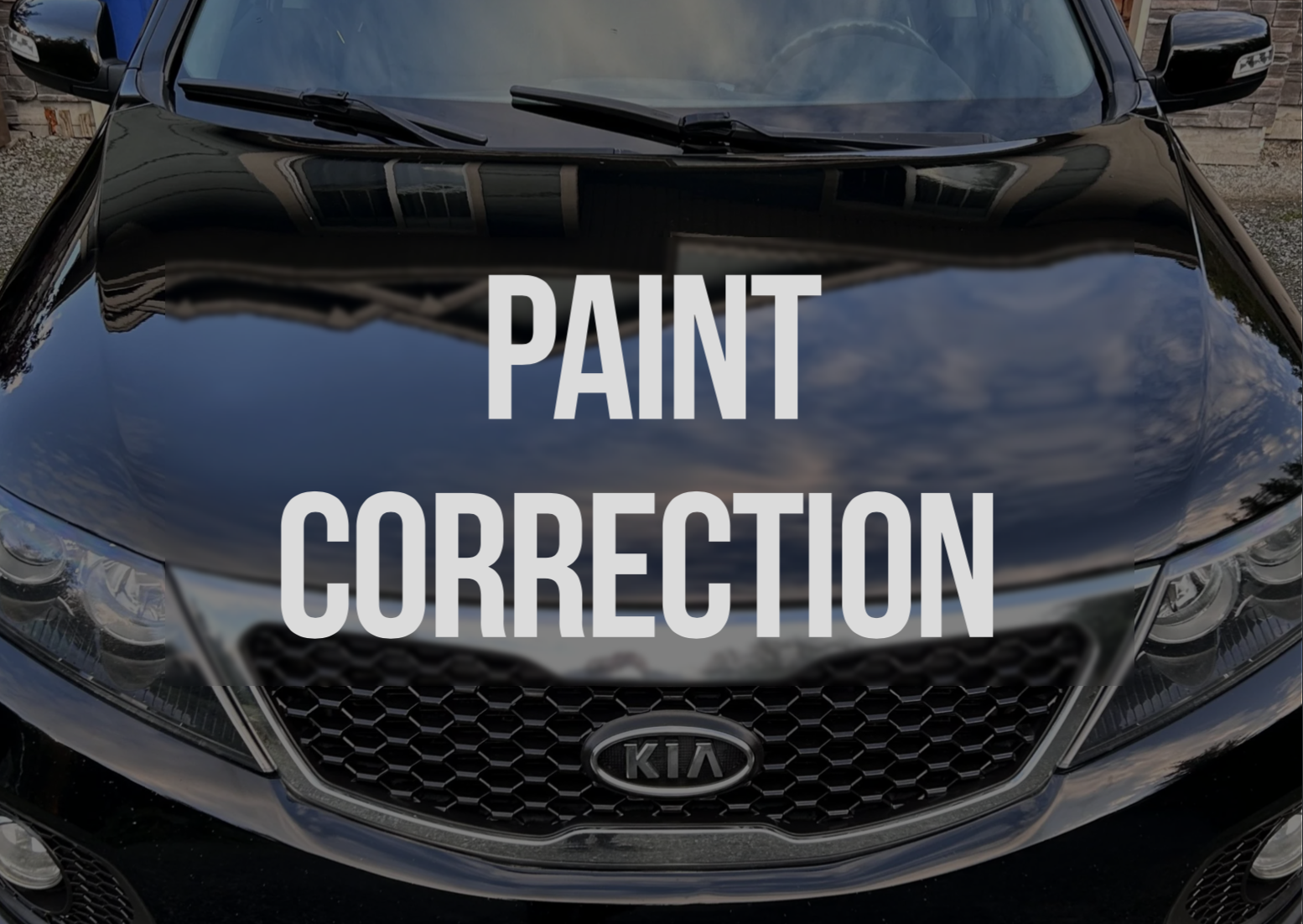 Front view of a black Kia car with the text 'Paint Correction' overlaid, showing the reflection of the sky and surrounding buildings on the car's hood.