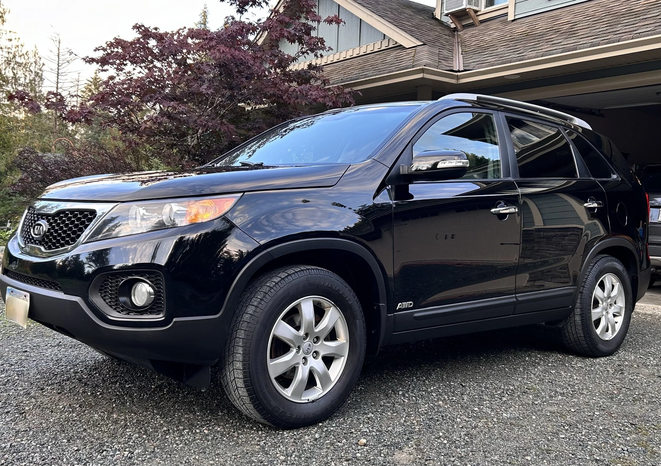 Black Kia SUV parked in a residential driveway with a house, trees, and a garage in the background.