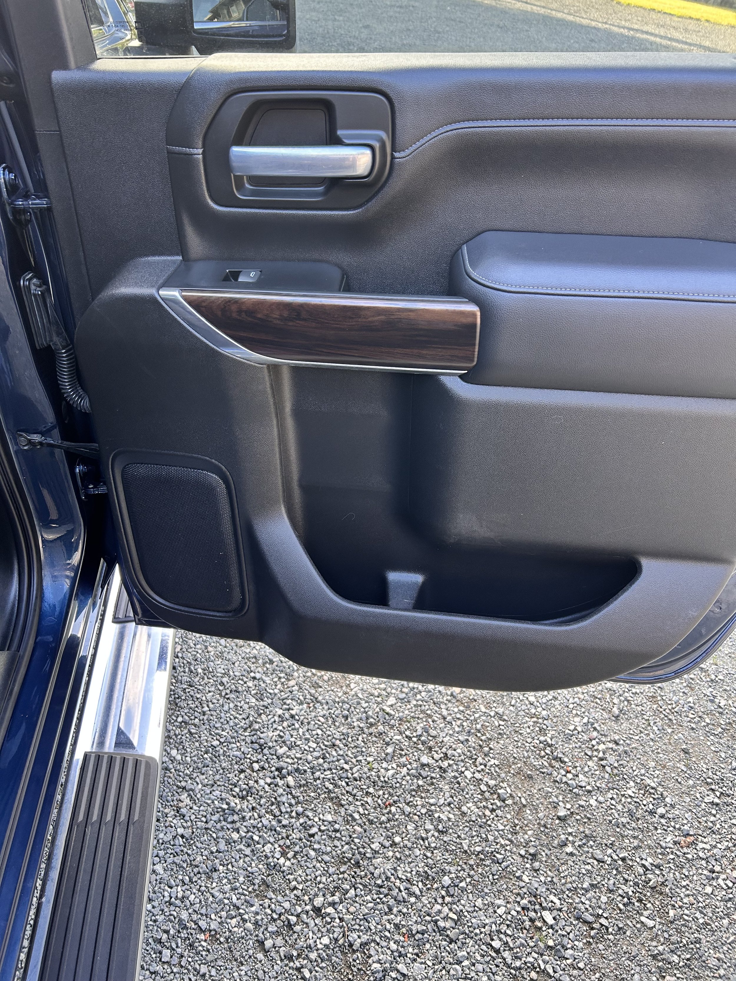 The interior of a vehicle door panel featuring a rubber handle, a window switch, a wood trim, and a storage compartment with a speaker at the bottom.