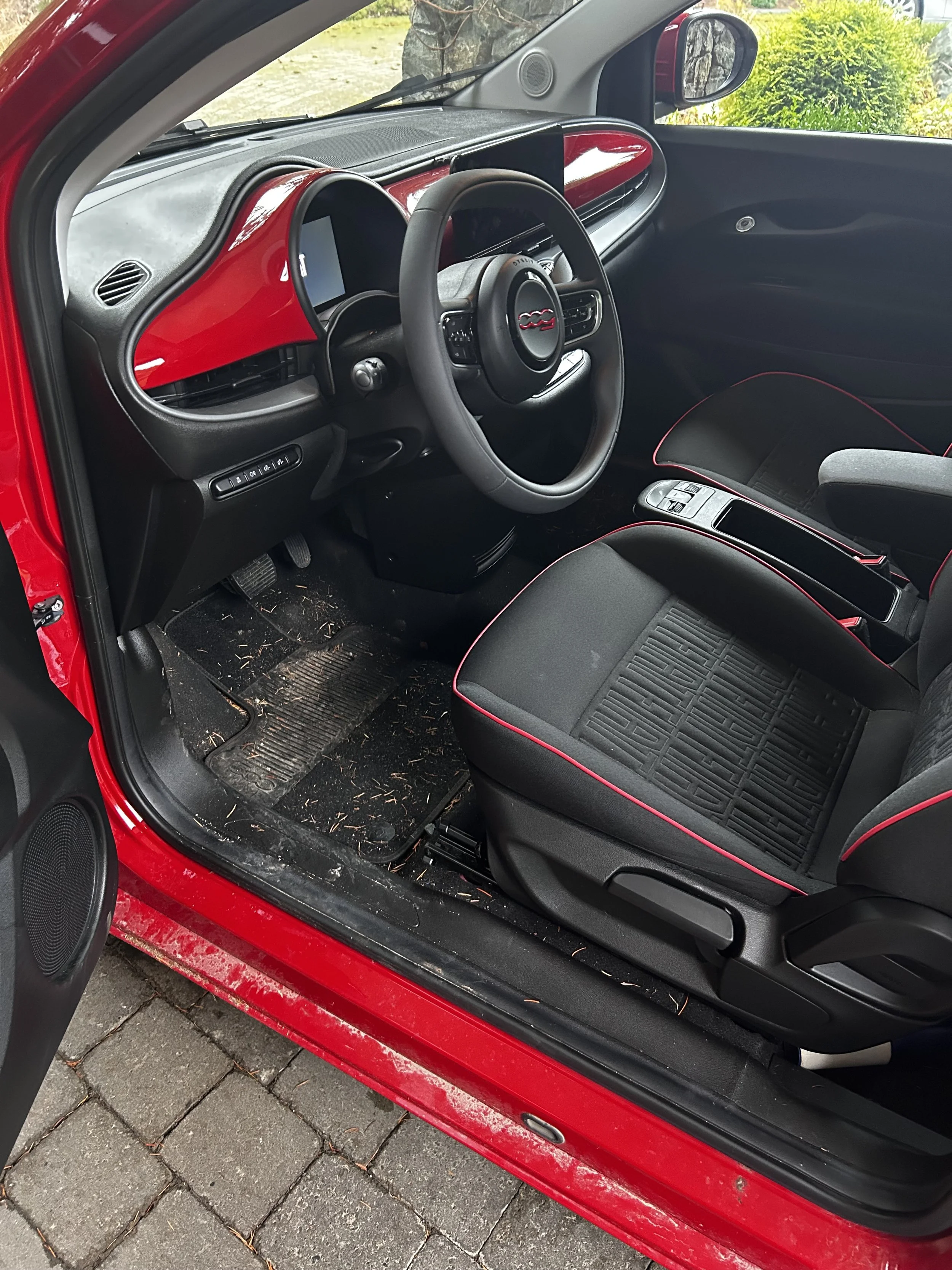 Interior of a red car showing the driver's seat, steering wheel, dashboard, and part of the front passenger seat with black seat covers with red piping, dirty car floor mats with debris.