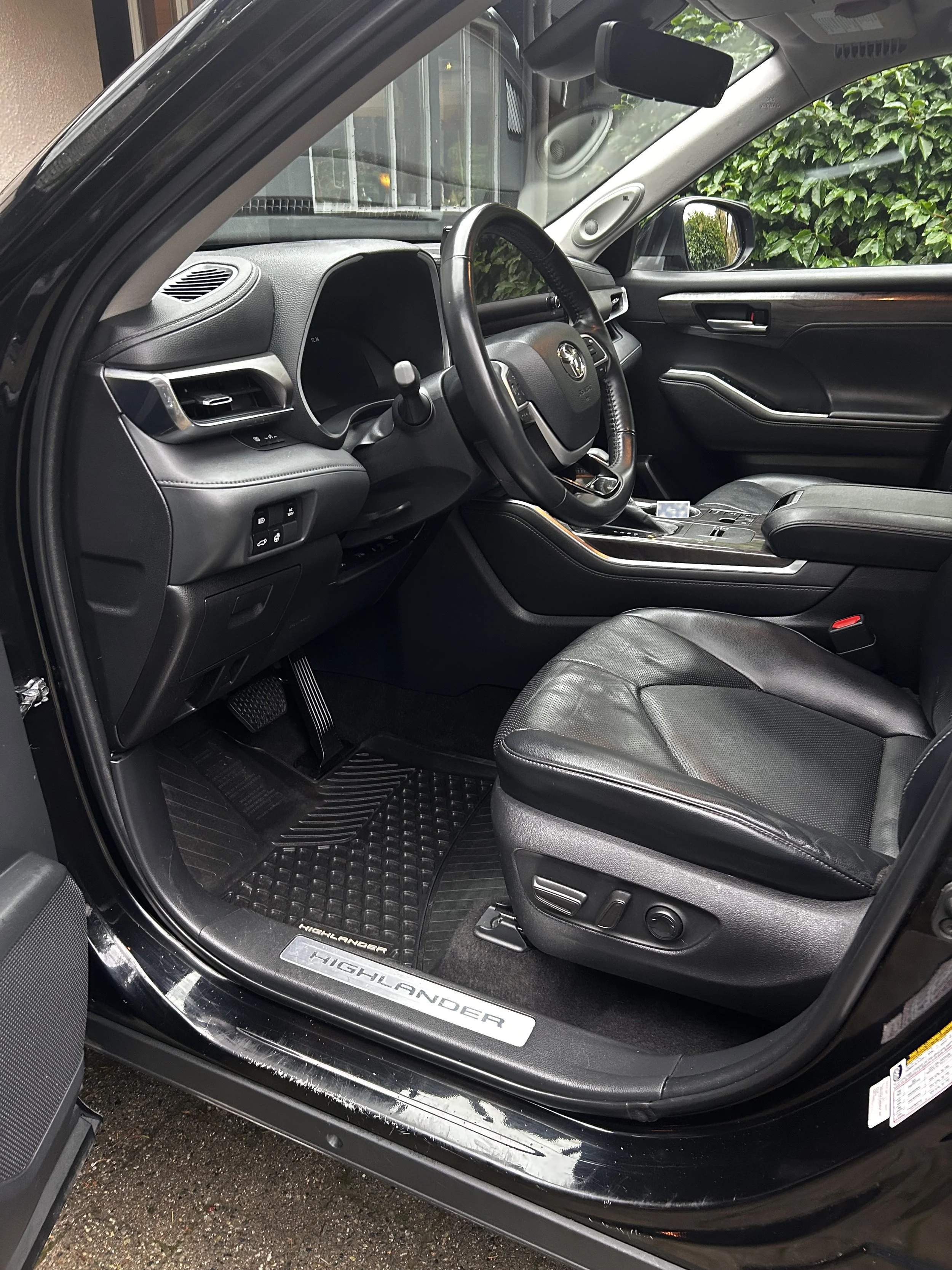 Interior of a Toyota Highlander SUV, featuring a black leather driver’s seat, dashboard, steering wheel, and center console, with green foliage outside the window.