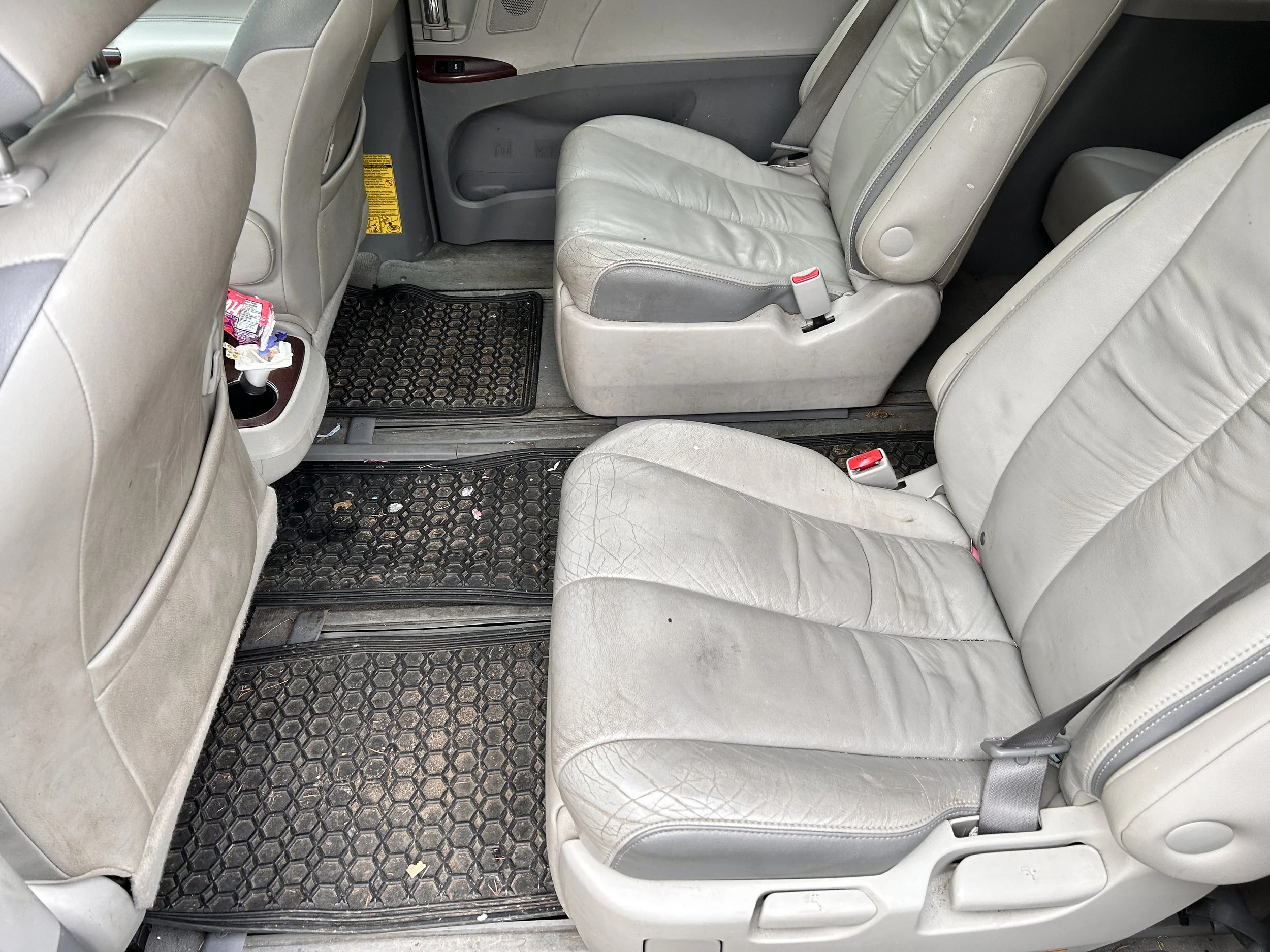 The interior of a car showing three rows of beige leather seats with black rubber floor mats, some trash and a cup holder with a snack wrapper.