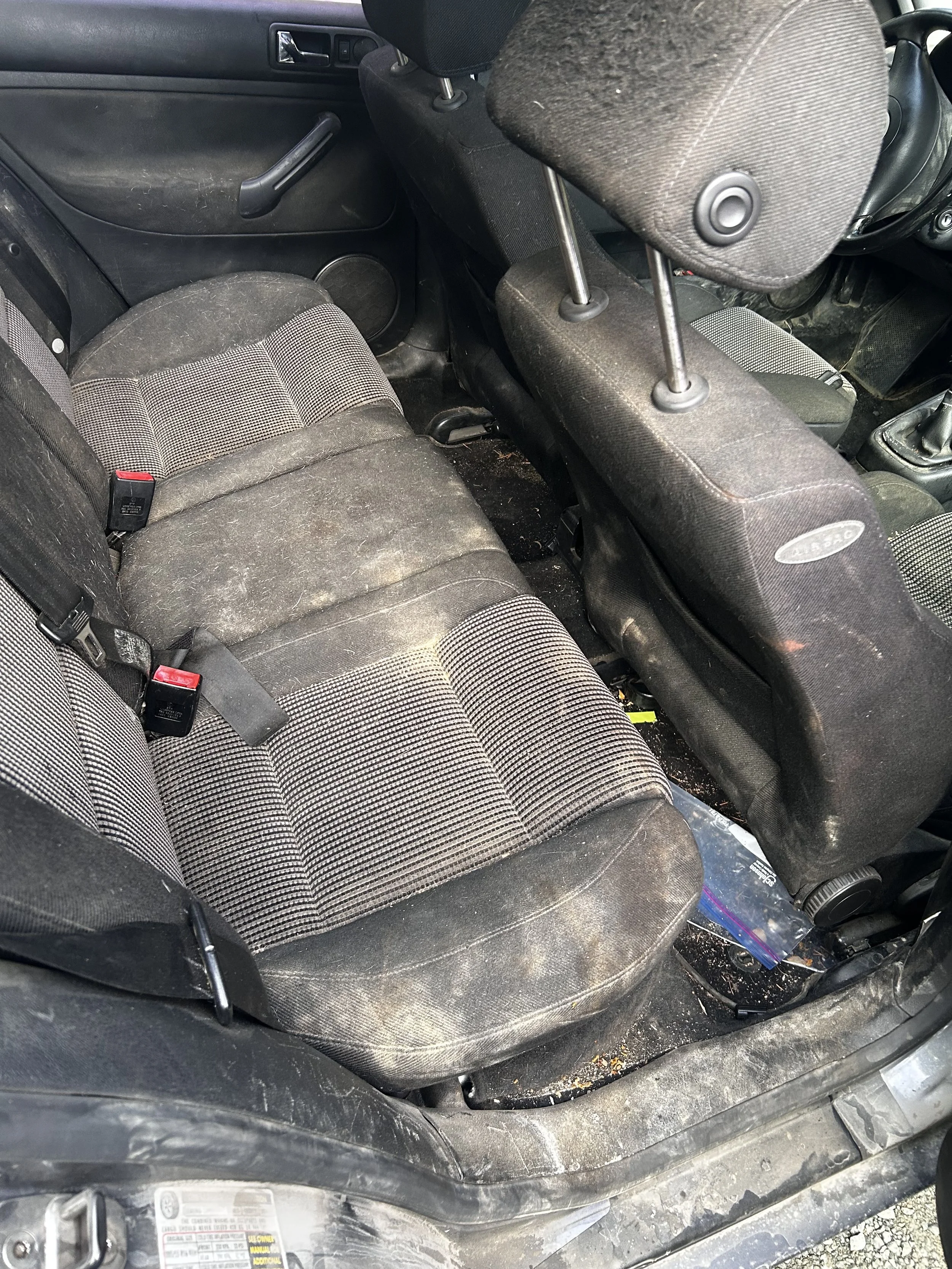 Interior of a car showing dirty, worn seats with fabric upholstery, a backseat with seat belts, and a dirty floor with debris.