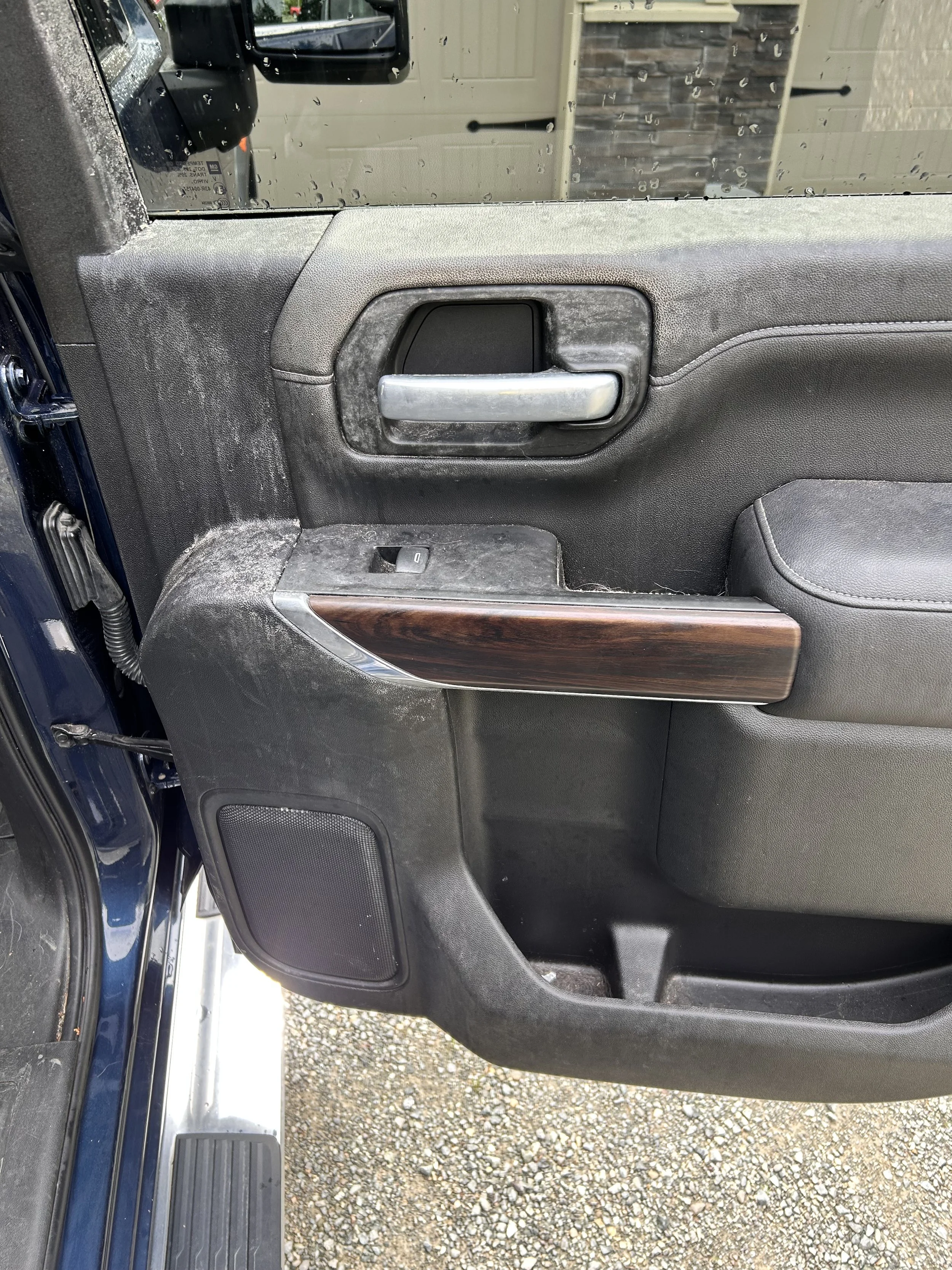 Close-up of a vehicle's interior door panel showing a silver door handle, a small window switch, a wood trim accent, and speaker, with dust and dirt present.