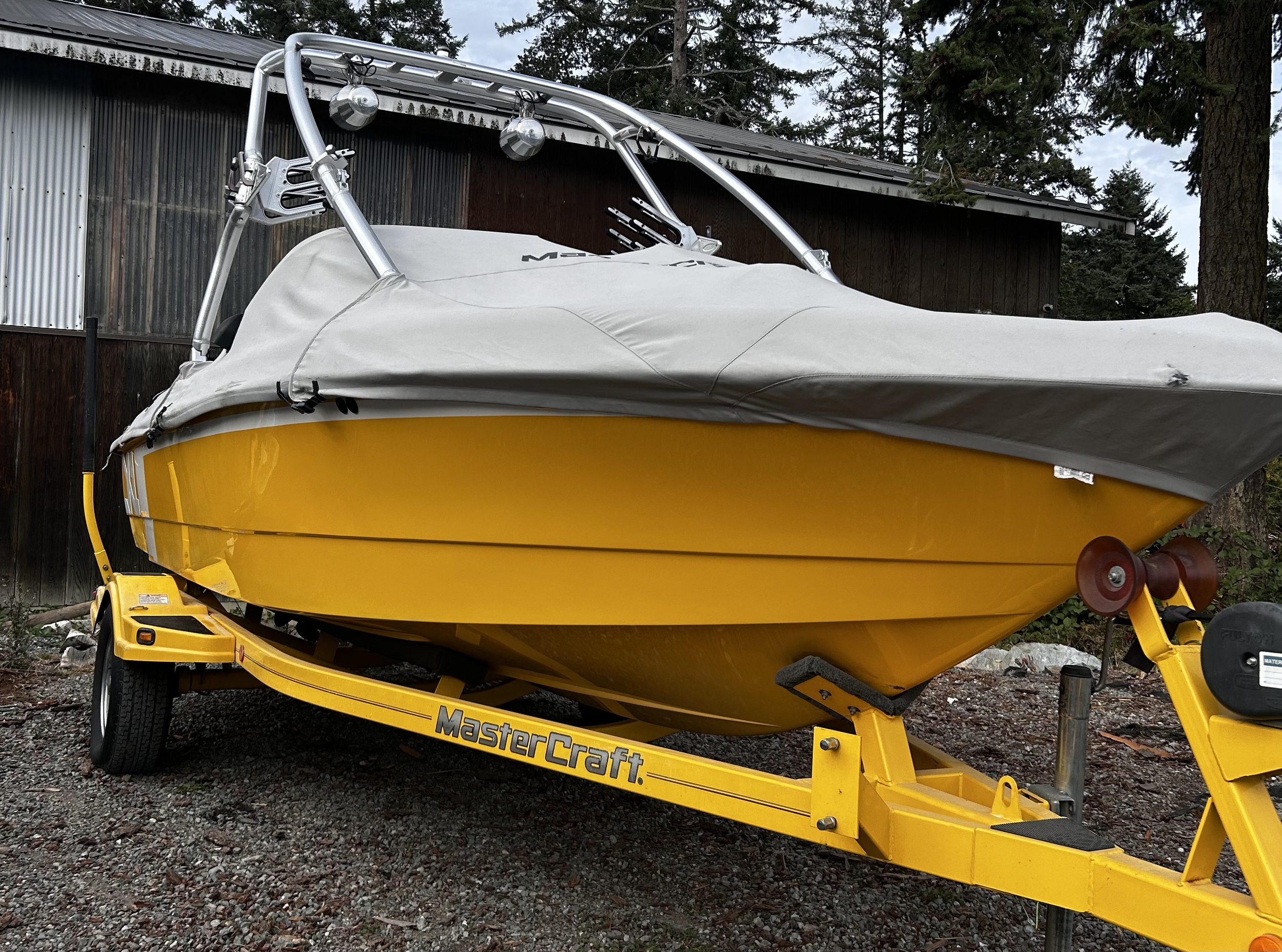 Yellow boat on a yellow MasterCraft trailer, covered with a gray cover, parked on a gravel surface near trees and a wooden building.