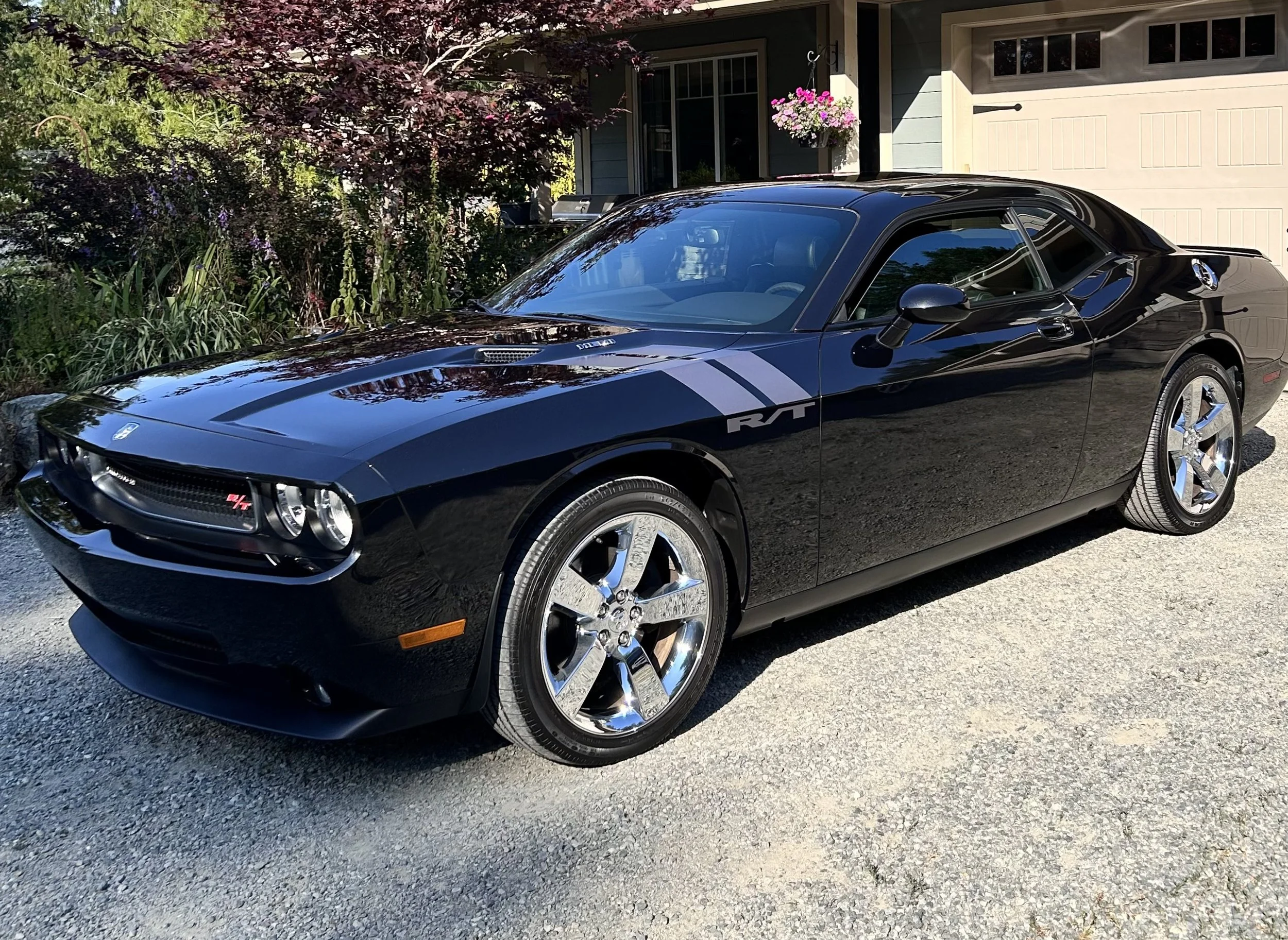 A black Dodge Challenger R/T parked on a driveway in front of a house with a garden. The car features a sleek design with chrome wheels, tinted windows, and a racing stripe on the side.