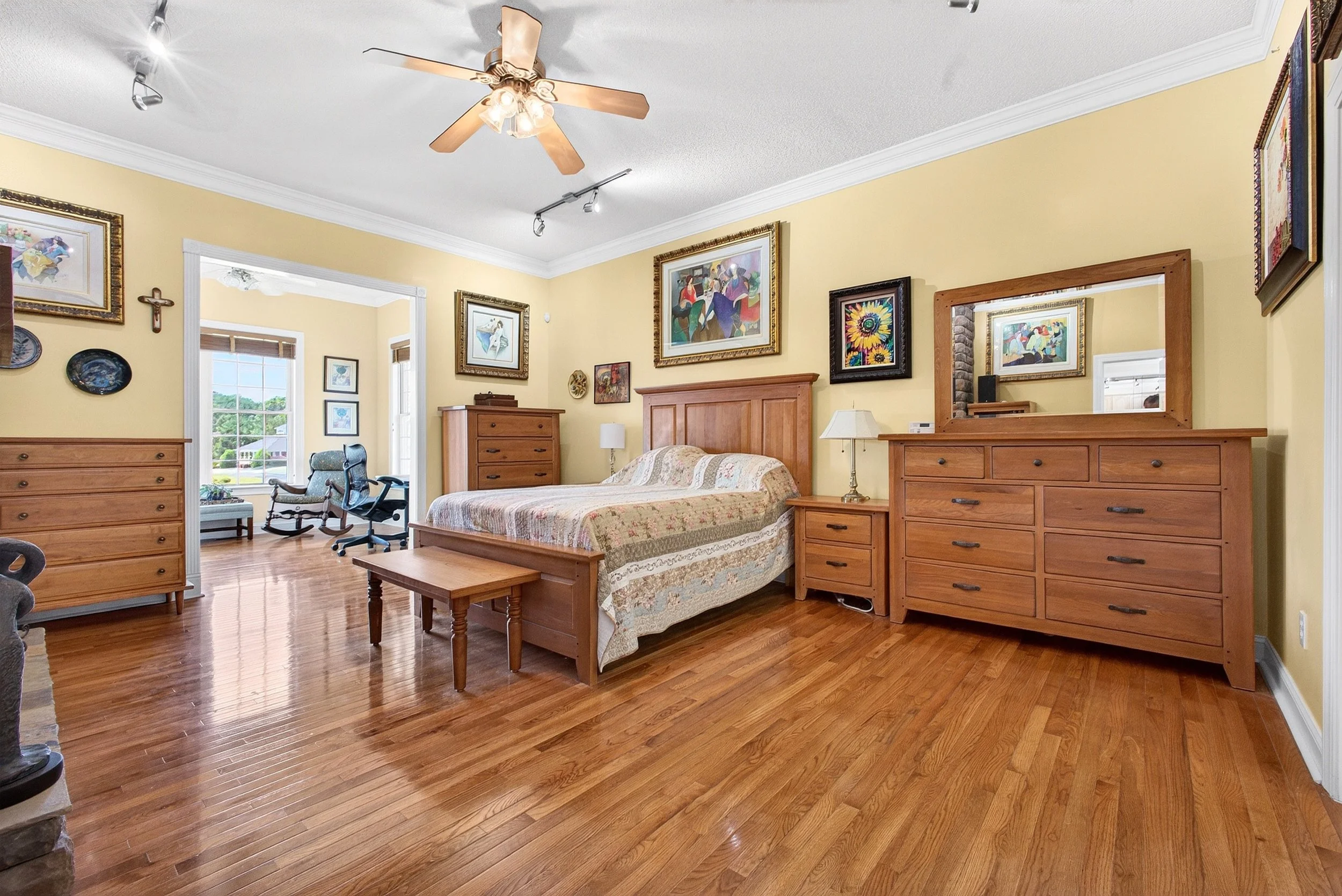 A spacious bedroom with yellow walls, wooden furniture including a bed, dressers, and a mirror, hardwood floors, and artwork hanging on the walls. There is a rocking chair and an office chair near the window, which has a view of green trees and a hou